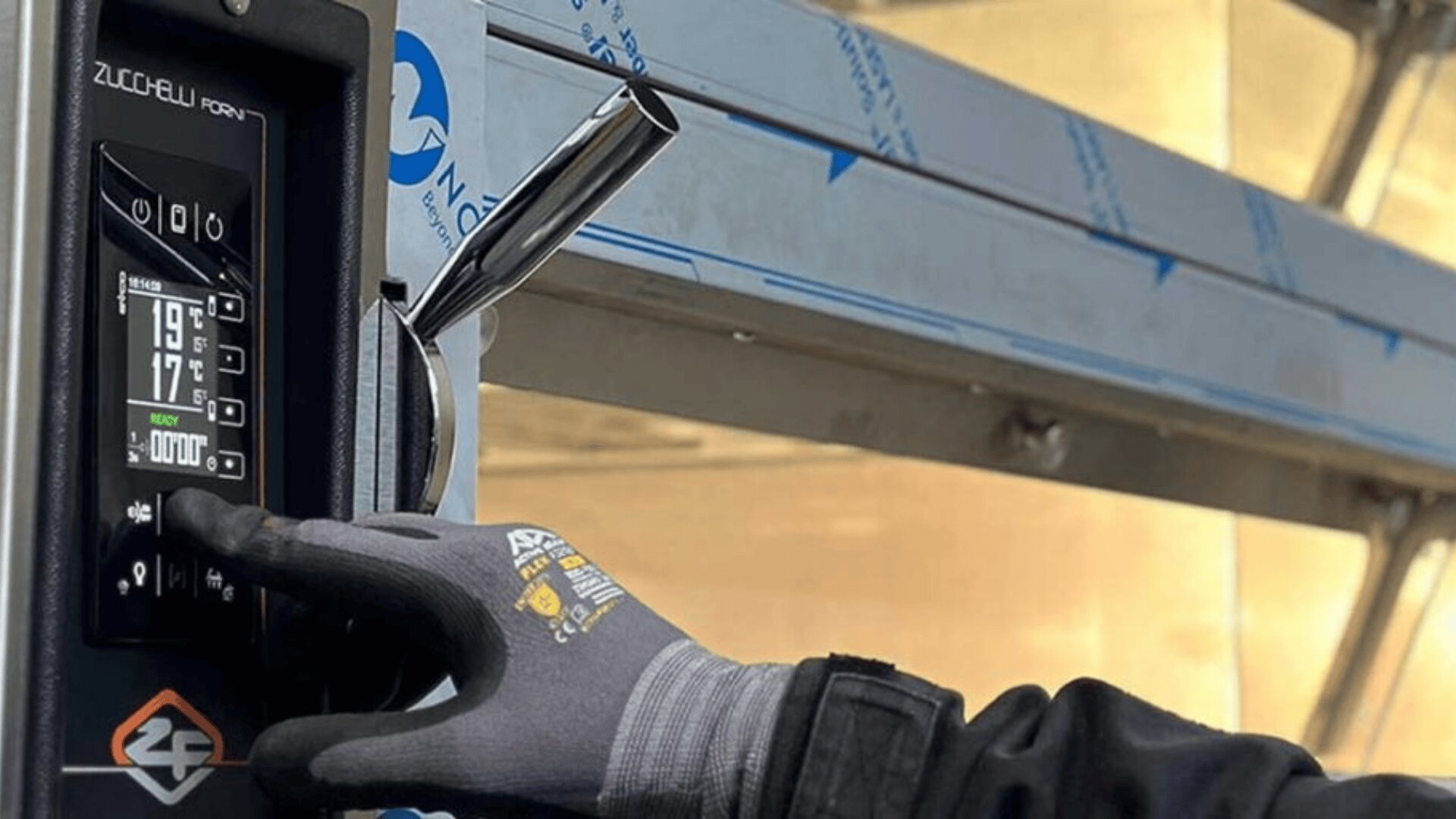 A person wearing a gray work glove is pressing buttons on a digital control panel of a machine, with metallic equipment and a blue-and-white logo visible in the background.