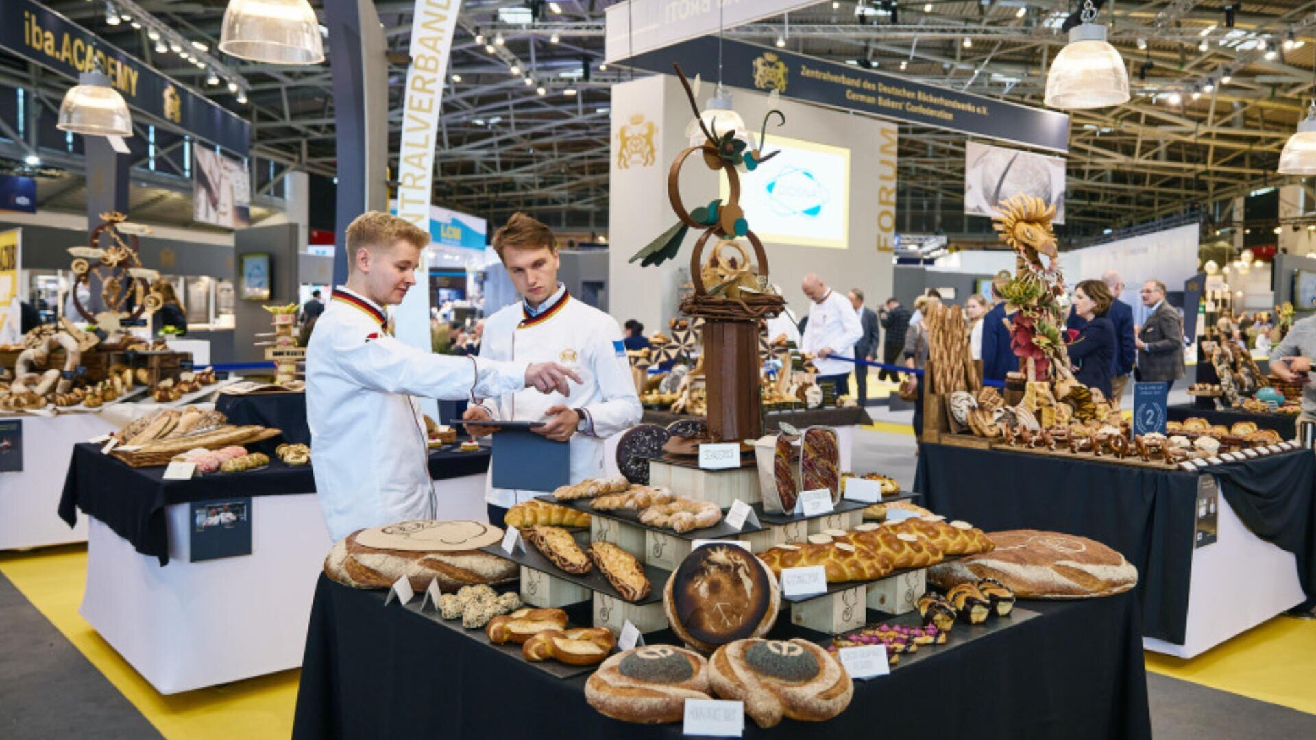 Zwei Männer in weißen Kochuniformen stehen bei einer Bäckereiausstellung an einem Auslagetisch mit verschiedenen handwerklich hergestellten Brotsorten und Gebäck, mit weiteren Brotauslagen und Menschen im Hintergrund.