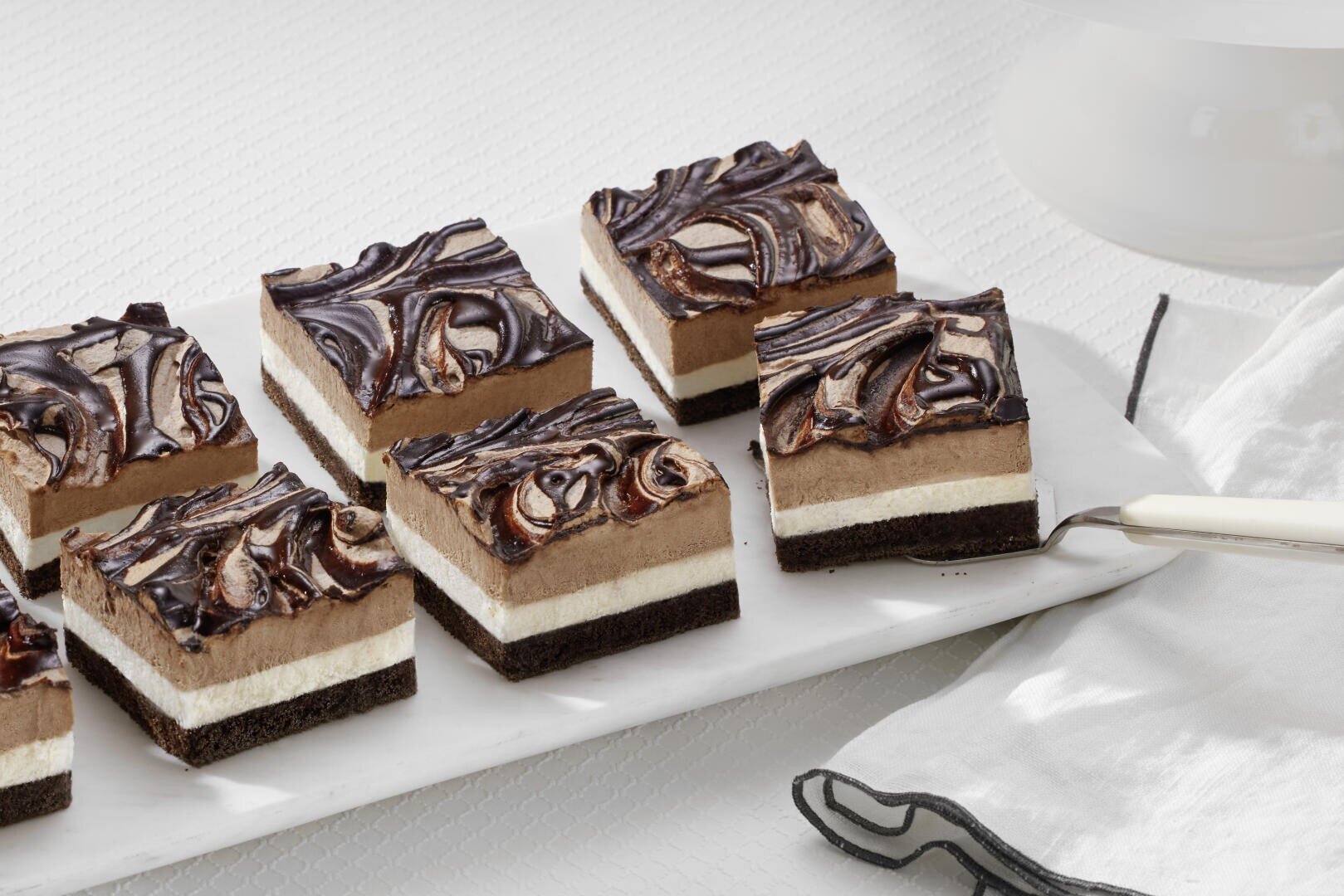 Neun quadratische Scheiben von geschichteten Schokoladen-Sahne-Dessertriegeln mit einem marmorierten Schokoladenüberzug sind auf einer weißen rechteckigen Platte neben einer Serviette und einem Servierbesteck auf einer weißen Fläche angeordnet.