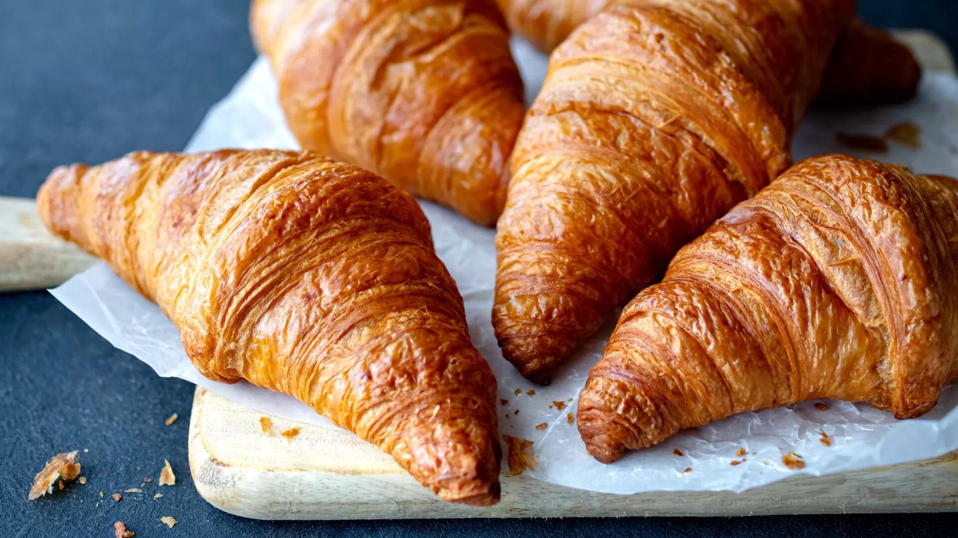 Vier goldbraune Croissants liegen auf Pergamentpapier auf einem Holzbrett. Das Gebäck ist flockig, mit sichtbaren Schichten und einer knusprigen Textur, und die Krümel sind auf einer dunklen Oberfläche verstreut.