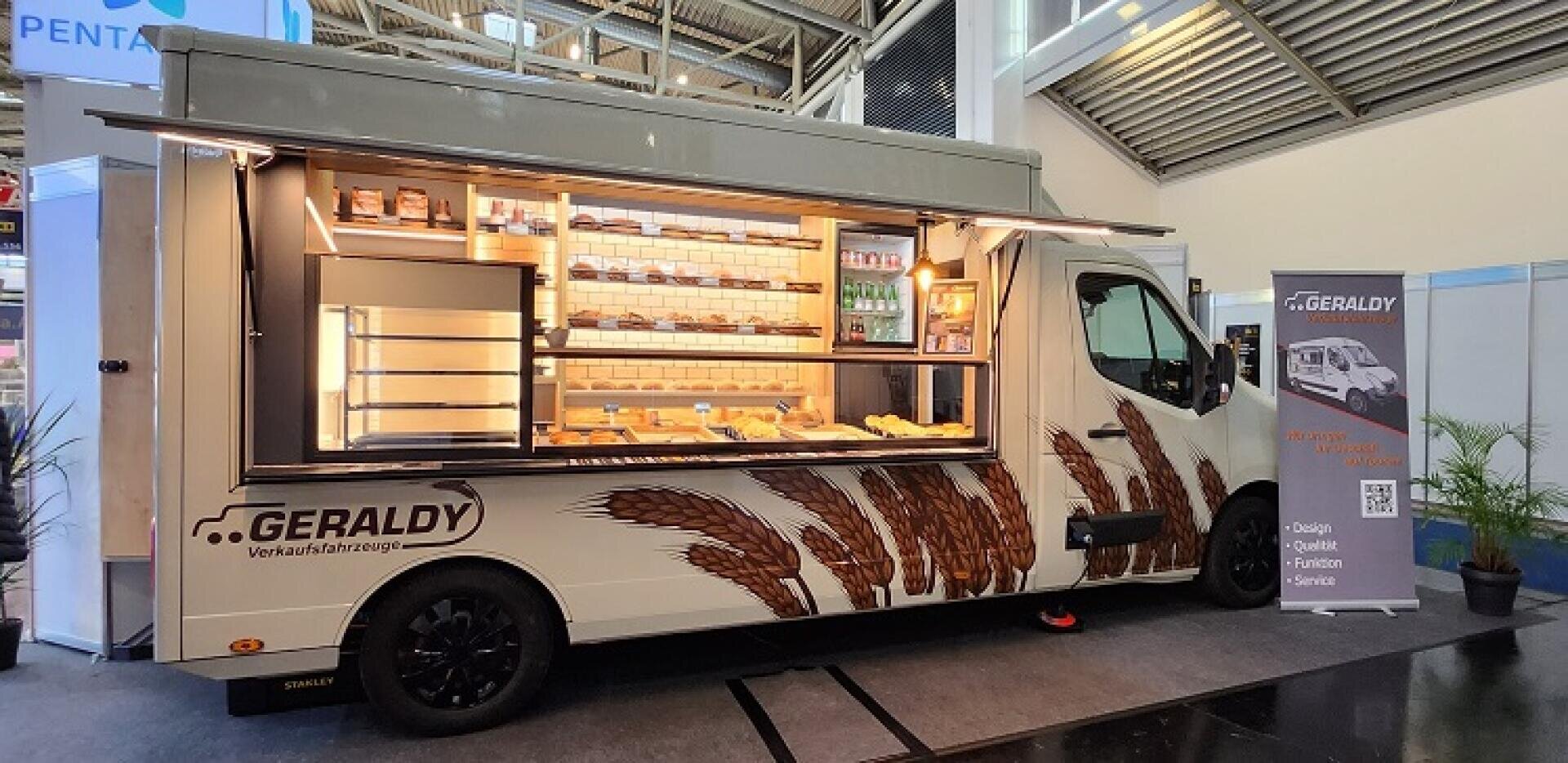 Ein mobiler Bäckerei-LKW mit offener Seite stellt Regale mit Brot und Gebäck aus. Der Wagen trägt die Marke GERALDY und ist mit Weizenmotiven versehen. Daneben befinden sich ein Poster und eine Topfpflanze in einem Innenbereich.