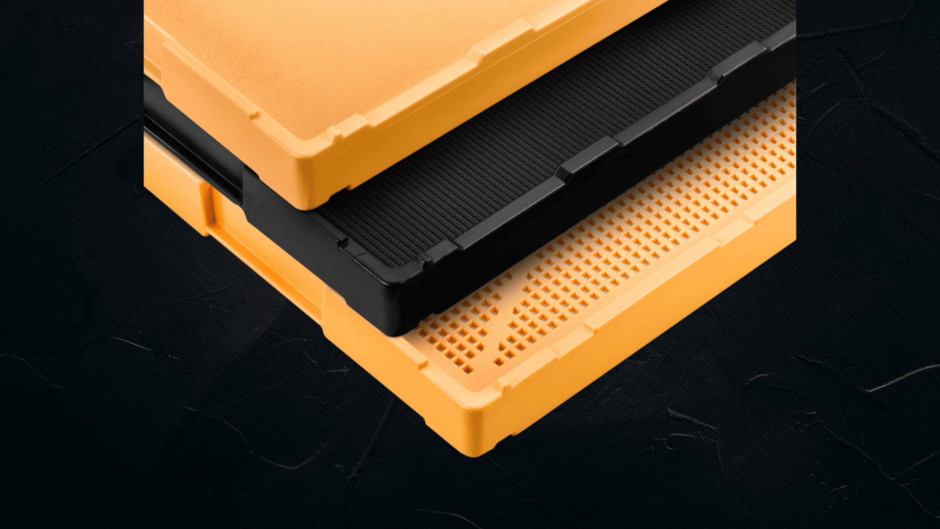 Nahaufnahme der Ecken von drei gestapelten Kunststoffschalen in Orange und Schwarz, wobei die obere Schale einfarbig, die mittlere schwarz und die untere Schale mit einem Lochgitter versehen ist. Der Hintergrund ist eine dunkel strukturierte Oberfläche.