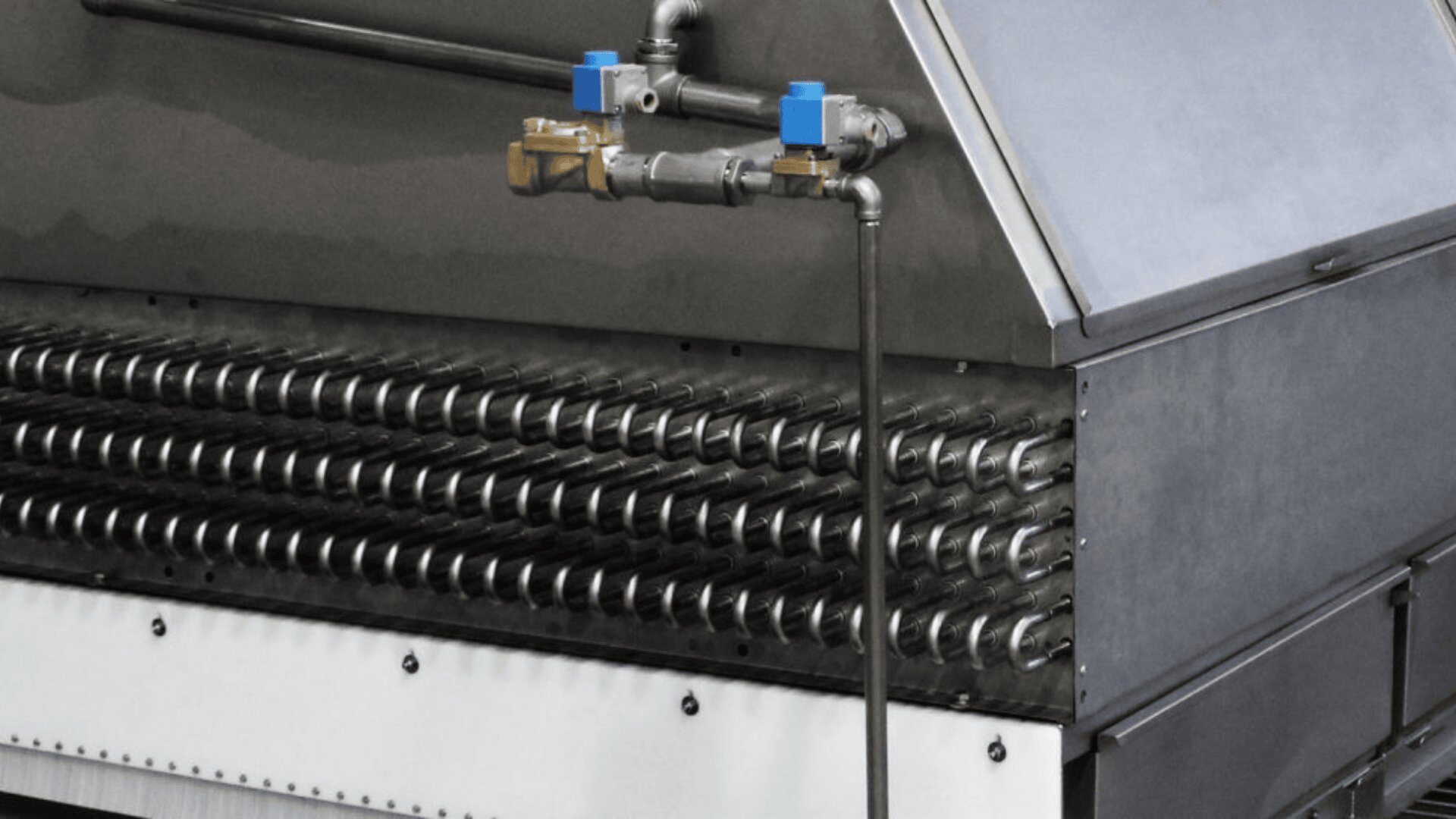 Nahaufnahme einer Industrieanlage mit mehreren Reihen horizontaler Metallrohre, Rohrleitungen mit blauen Ventilen darüber und einem großen Metallgehäuse auf der rechten Seite.