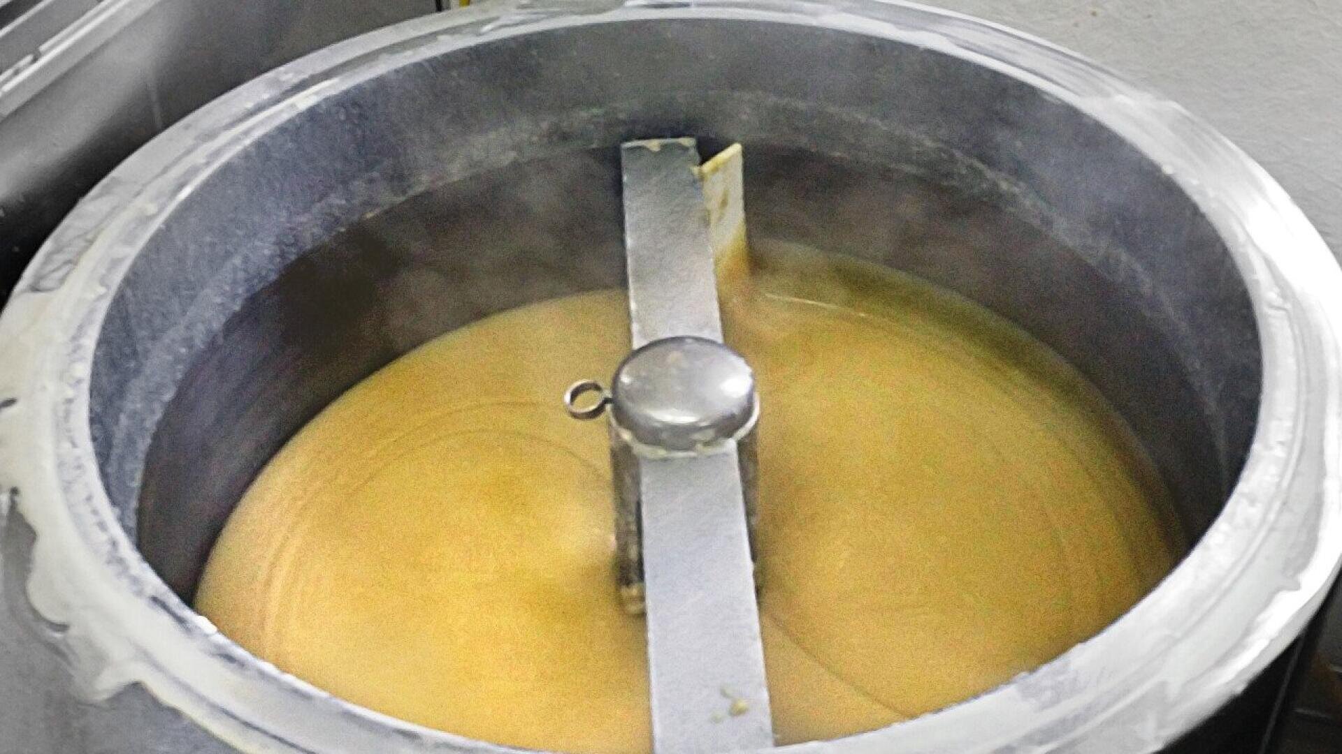 A large industrial metal pot filled with steaming, creamy yellow liquid being stirred by a metal paddle attached to a rotating arm.