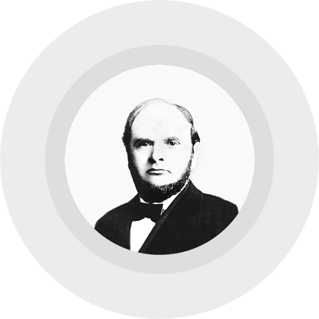 Schwarz-Weiß-Porträt eines Mannes mit kurzen Haaren, Koteletten und einem Bart. Er trägt einen Anzug mit Fliege vor einem schlichten hellgrauen Hintergrund in einem runden Rahmen.