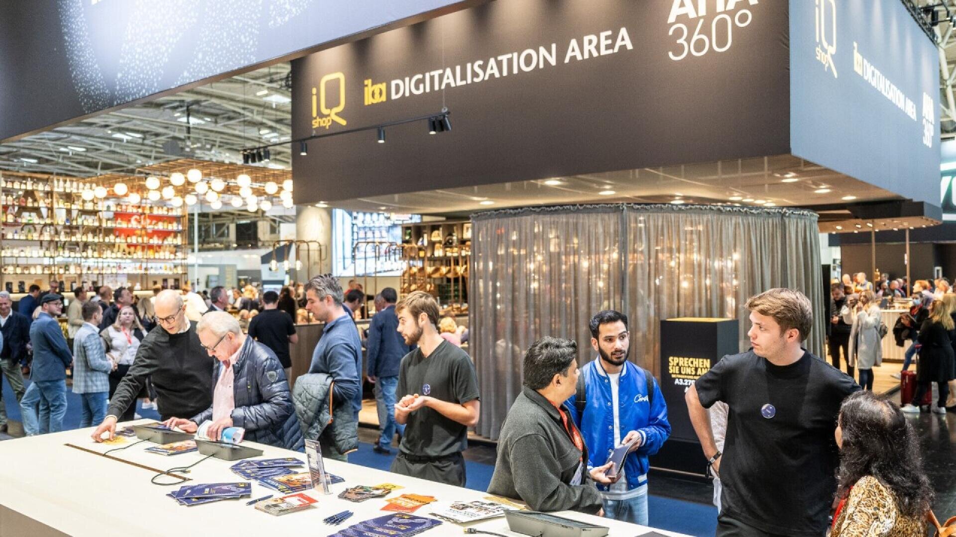 A busy exhibition hall features a “Digitalisation Area” booth, with several people interacting at counters and talking. The background shows a crowded event space with lights and displays.
