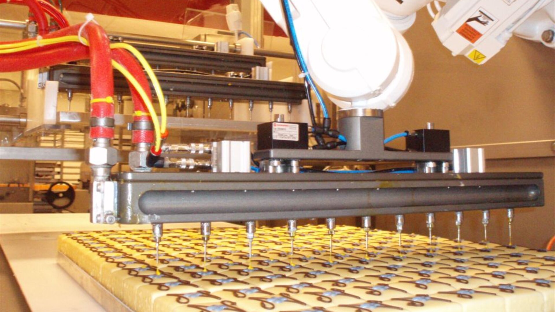 A robotic arm decorates rectangular pastries with chocolate swirls on a production line in a food processing facility. Red and yellow tubes supply ingredients to the machine.
