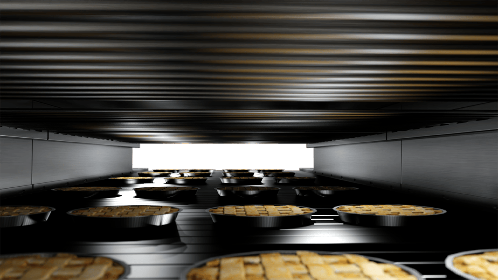Rows of small lattice-topped pies on a conveyor belt, baking inside a large industrial oven, viewed from a low angle.