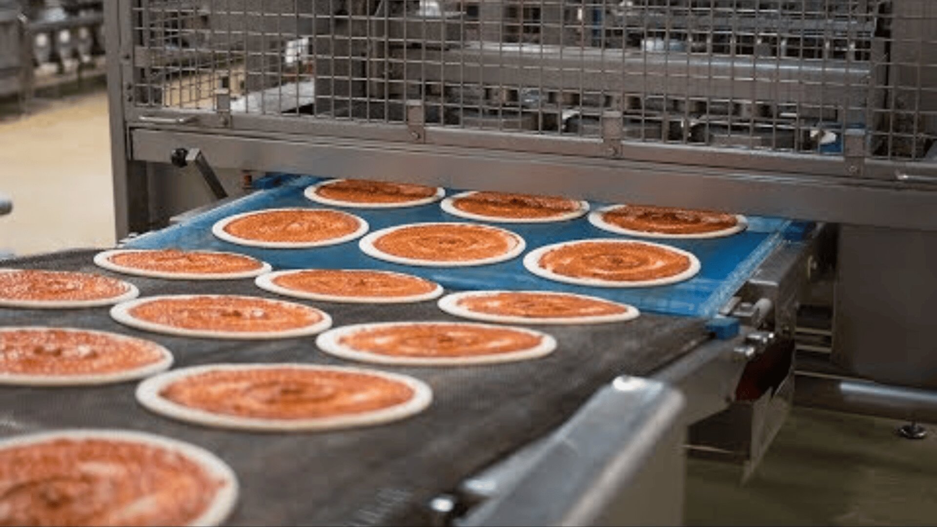 Several raw pizzas with tomato sauce move on a conveyor belt in an industrial food production plant, with metal machines visible in the background.