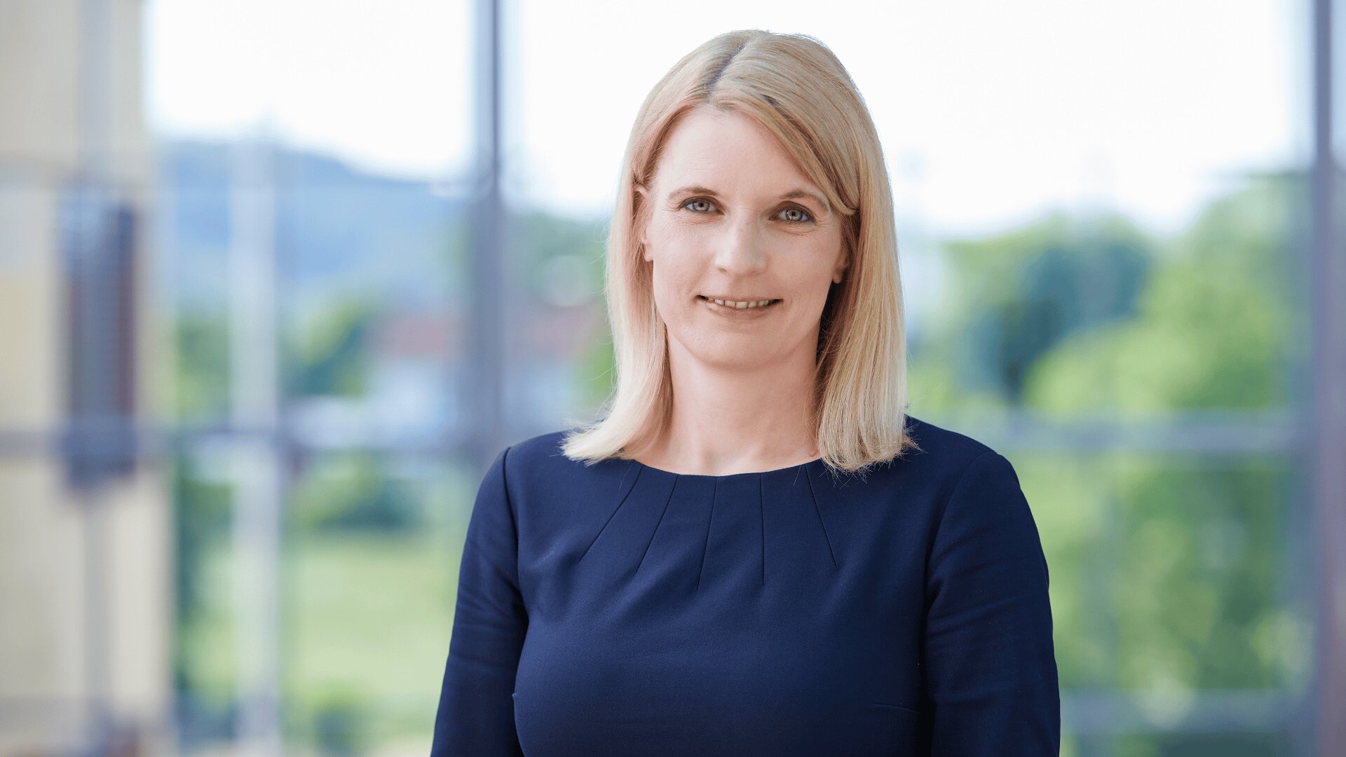 Eine Frau mit glattem blondem Haar und einem marineblauen Kleid steht in einem Innenraum vor großen Fenstern, im Hintergrund sind Grünpflanzen und eine unscharfe Außenszene zu sehen. Sie lächelt sanft in die Kamera.