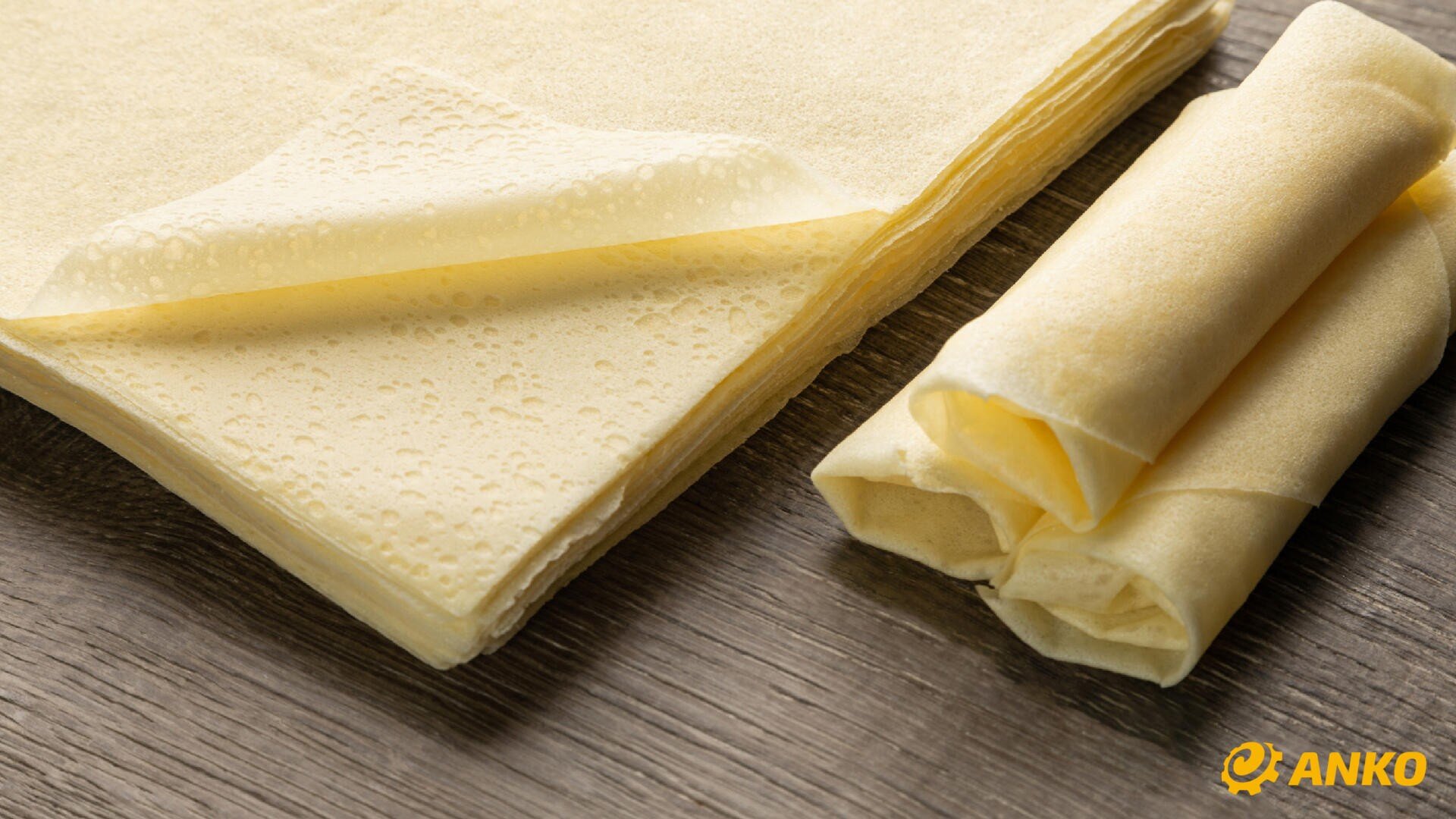 Thin, rectangular sheets of yellowish pastry lie on a wooden surface, partly stacked and partly rolled. The ANKO logo can be seen in the bottom right-hand corner.