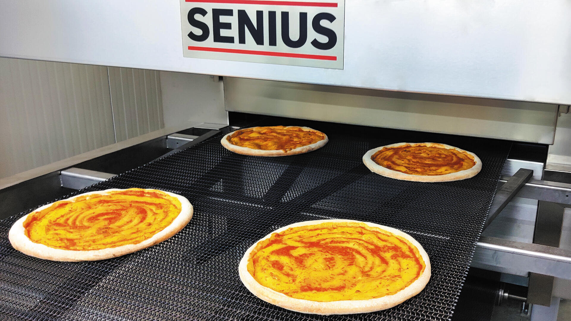 Vier rohe Pizzaböden mit Tomatensauce laufen über ein Förderband in einen Industrieofen mit der Aufschrift SENIUS in einer Lebensmittelproduktionsstätte.