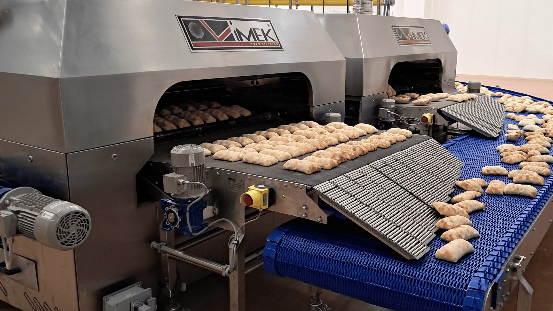 Conveyor belts with pastries move through large industrial baking machines in a food processing facility, with freshly baked goods exiting onto blue and black conveyor trays.