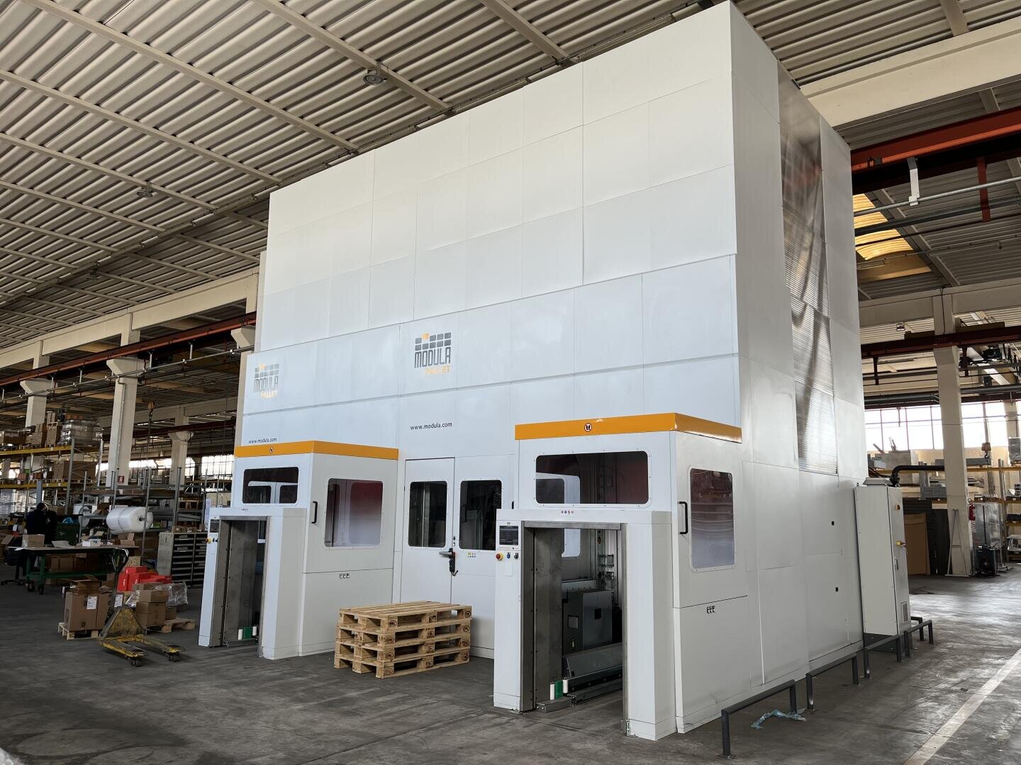 Large industrial machine with multiple doors and windows, situated inside a spacious warehouse with a high ceiling. Wooden pallets and boxes are nearby, and other machinery is visible in the background.