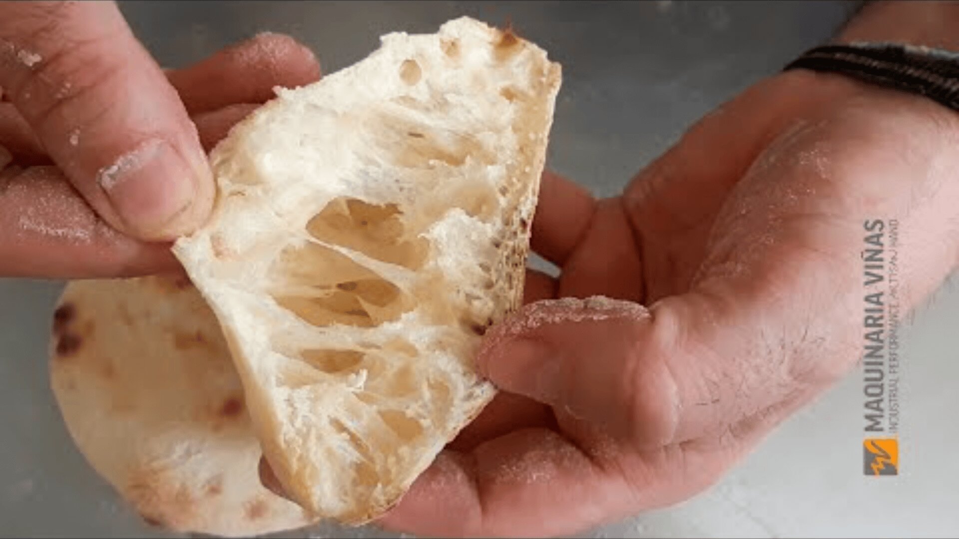 A close-up of hands holding a piece of rustic, airy and holey bread, showing off its open crumb structure. The bread appears to be fresh and lightly dusted with flour. The logo of Maquinaria Viñas can be seen on the right.