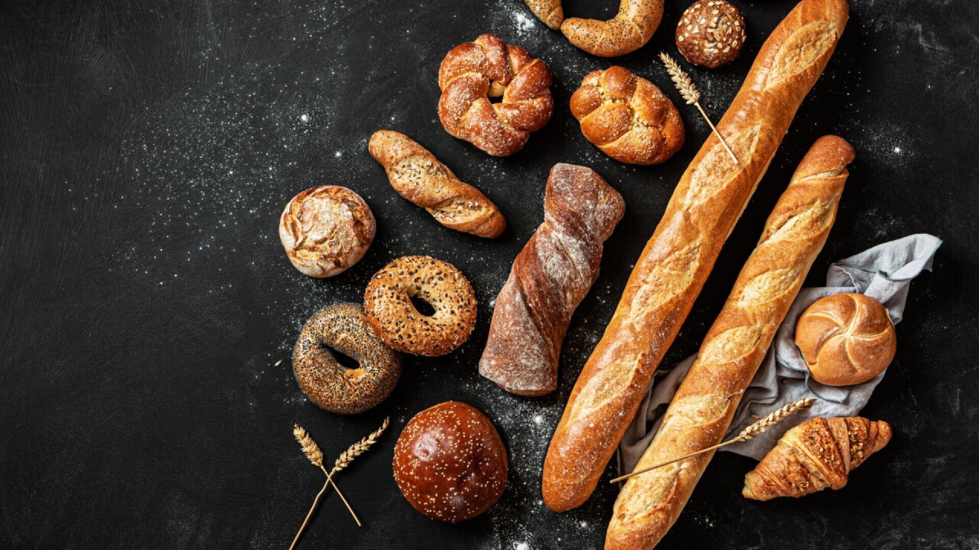 An assortment of various breads, including baguettes, bagels, rolls, croissant, and rustic loaves, arranged on a dark surface with scattered flour and wheat stalks.