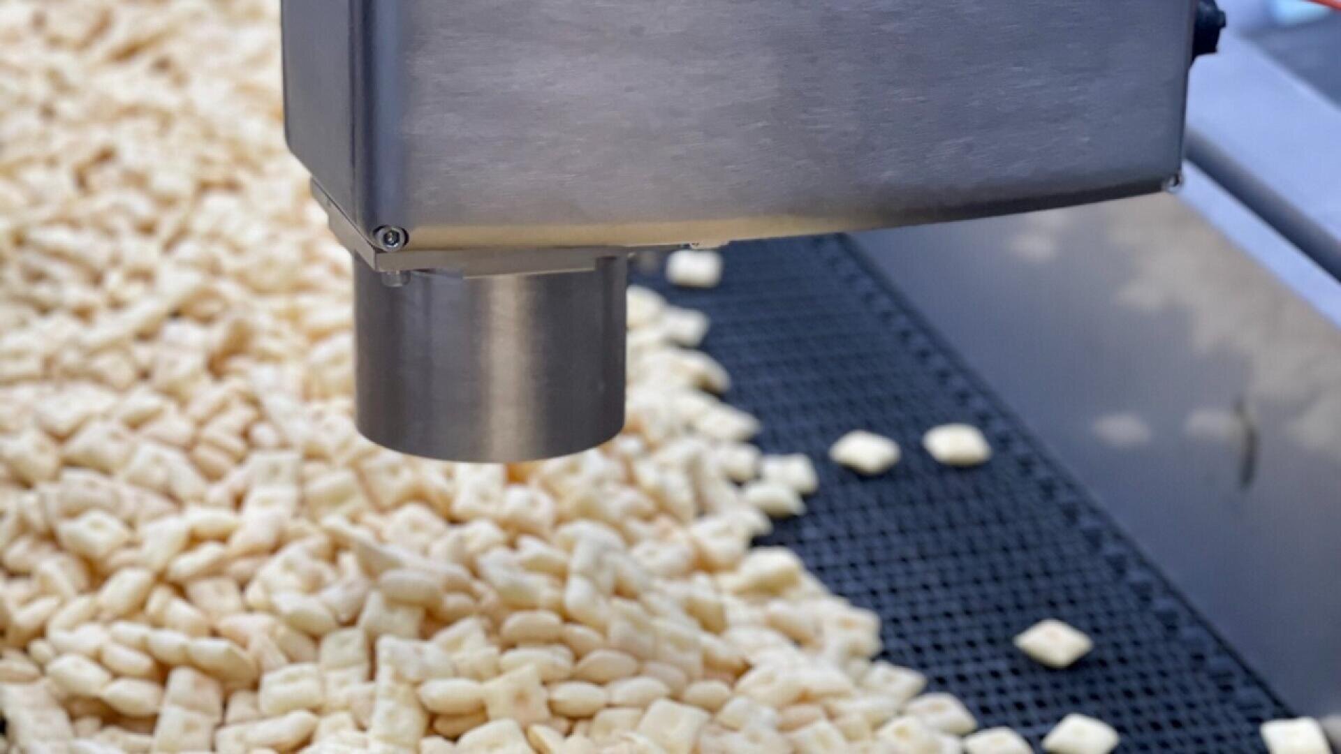 Eine Nahaufnahme einer Lebensmittelverarbeitungsmaschine, die kleine quadratische Getreidestücke auf ein Förderband in einer Fabrikumgebung abgibt.