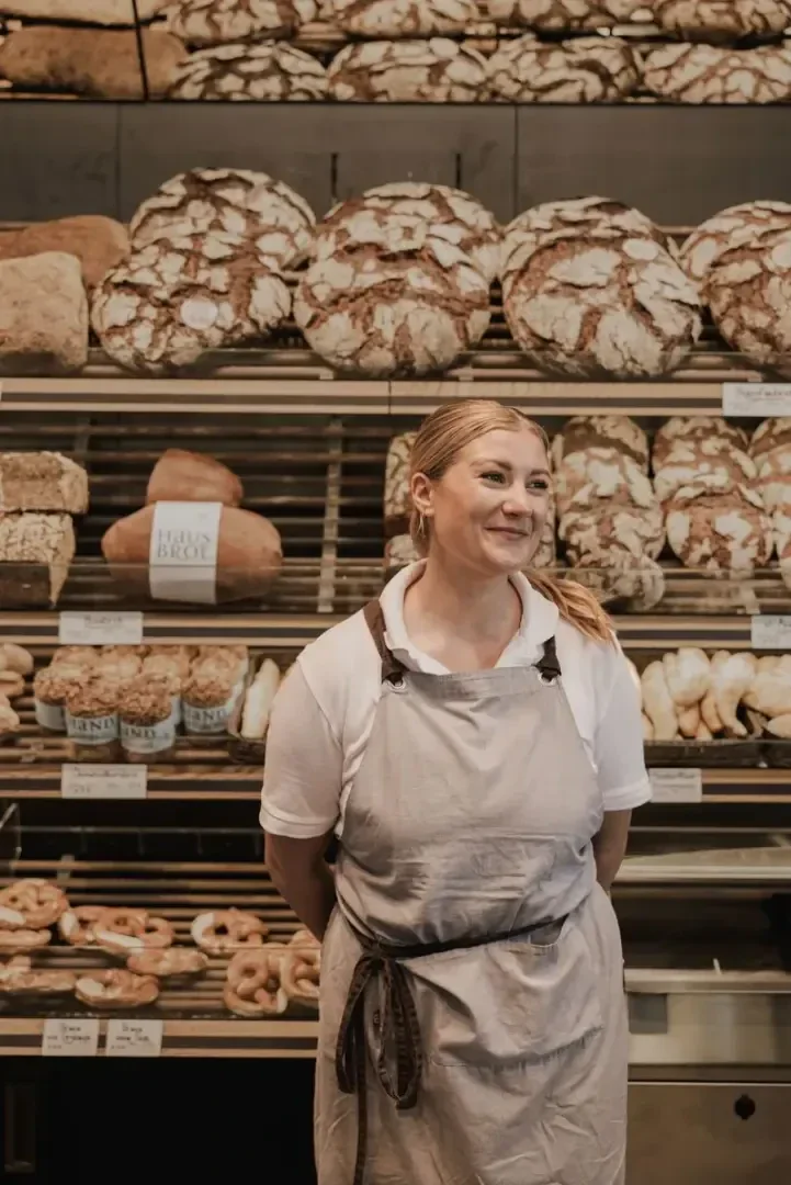 Eine Frau mit grauer Schürze und weißem Hemd steht lächelnd vor Regalen voller Brot und Backwaren in einer Bäckerei.