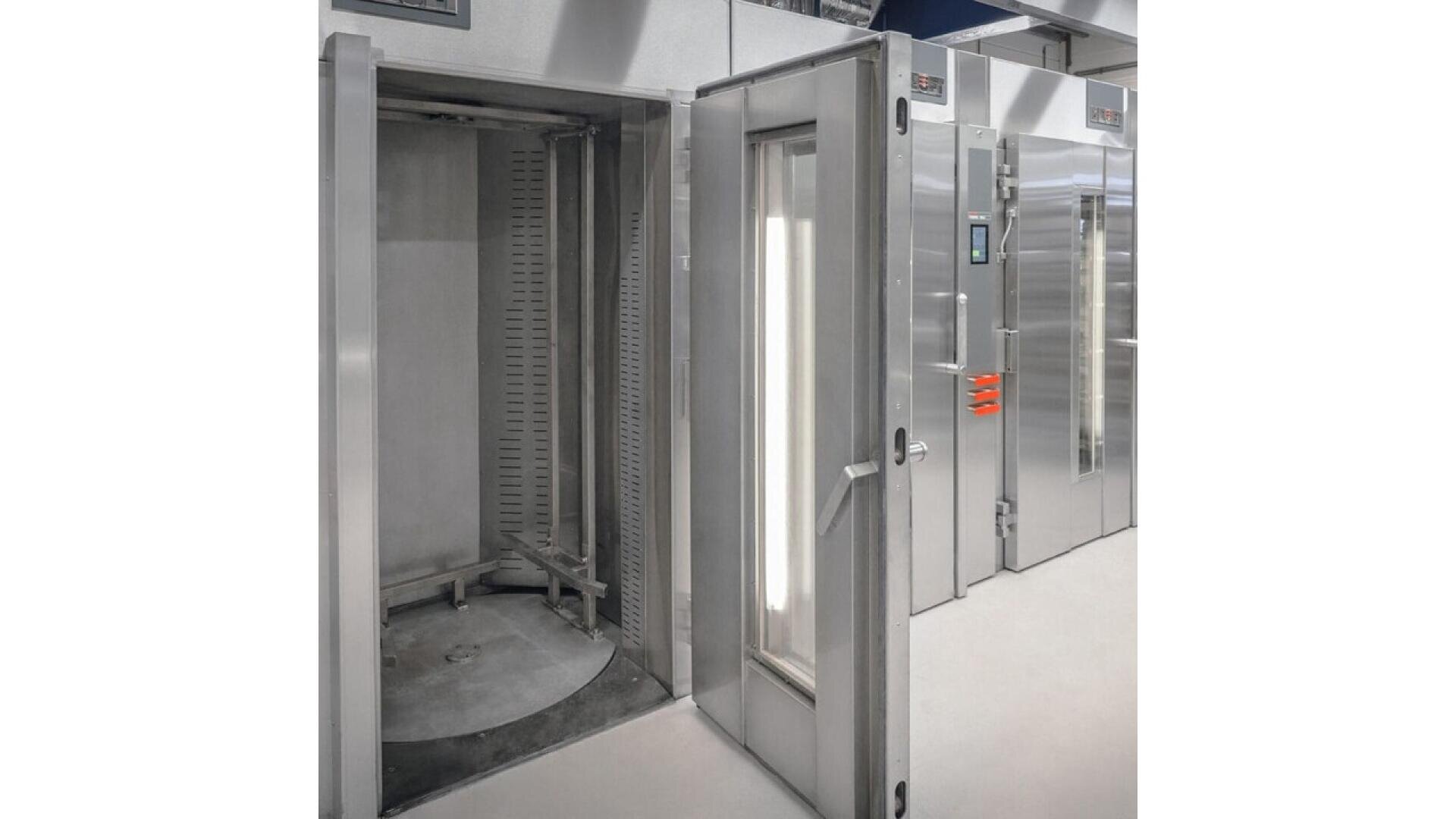 A large industrial stainless steel oven with an open glass door, revealing a circular rotating tray and a clean, modern interior. Additional closed ovens are visible beside it in a bright, sterile environment.