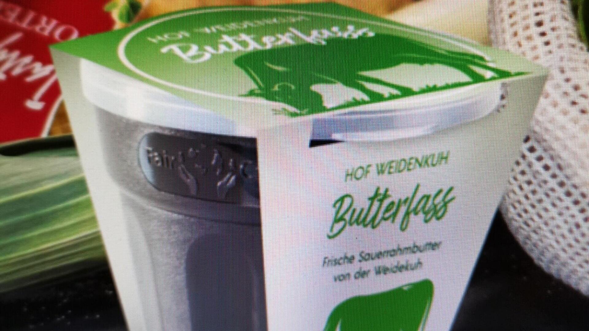 A container of “Hof Weidekuh Butterfass” fresh sour cream, featuring green and white packaging with a cow graphic and German text, placed on a table near vegetables.