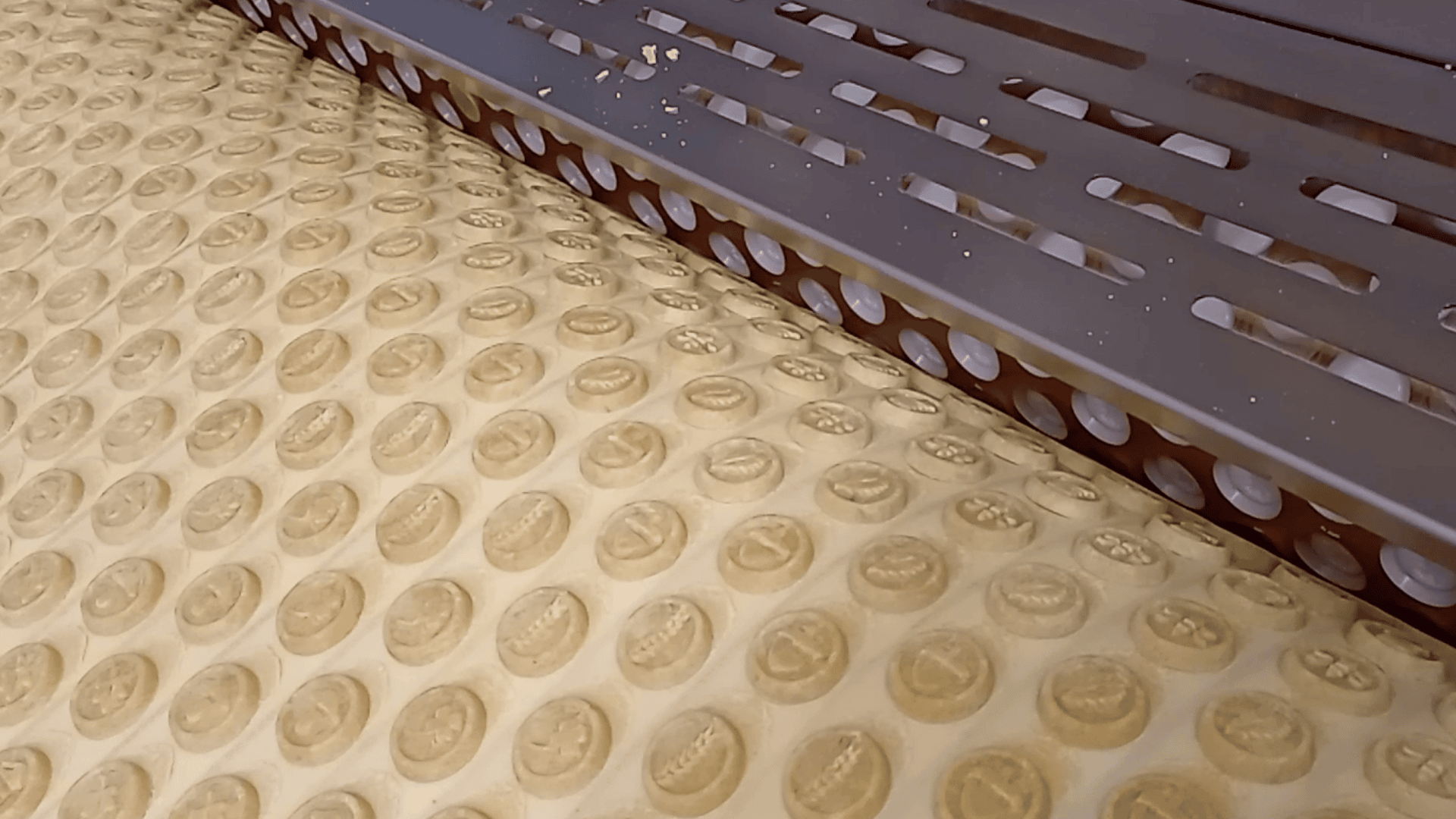 A machine presses rows of round biscuits with designs onto a large sheet of dough, cutting and shaping them for production. Some crumbs are visible near the cutting edge.