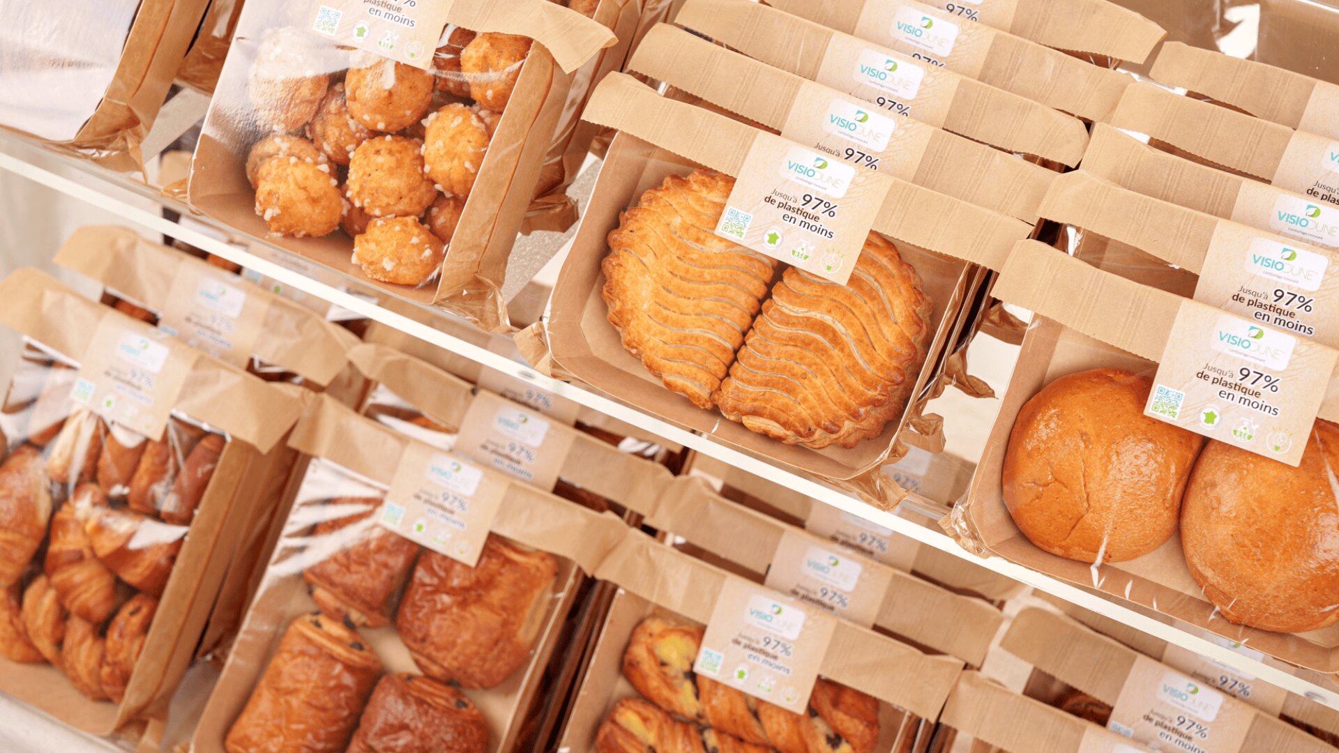 Assorted baked goods, such as pastries and bread rolls, are displayed on shelves in paper packaging with labels. The labels show information about the products’ ingredients and nutritional content.