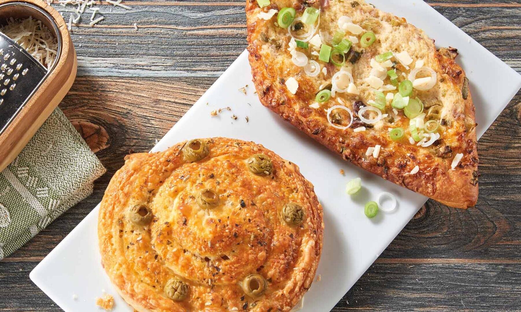 Ein Käsestrudel mit grünen Oliven und eine Scheibe Knoblauchbrot mit geschmolzenem Käse und grünen Zwiebeln auf einem weißen Teller, der auf einem Holztisch steht.