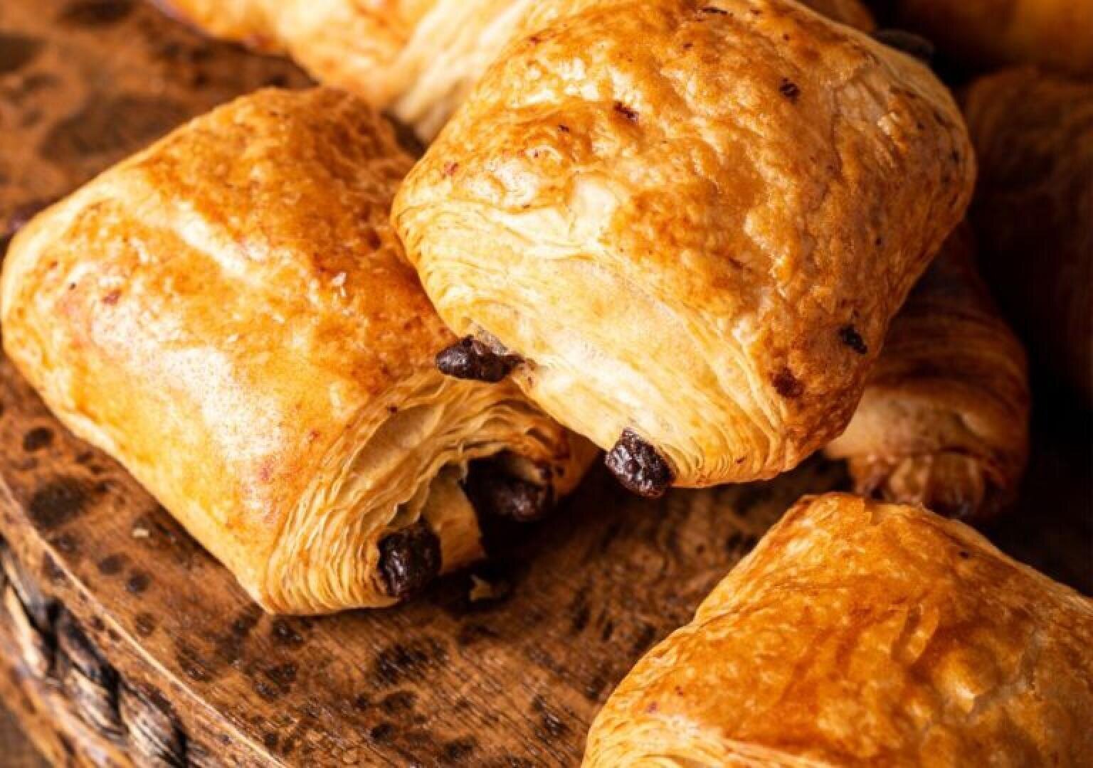 Several golden-brown pain au chocolat pastries rest on a rustic wooden surface, showing their flaky, layered texture and peeking chocolate filling.