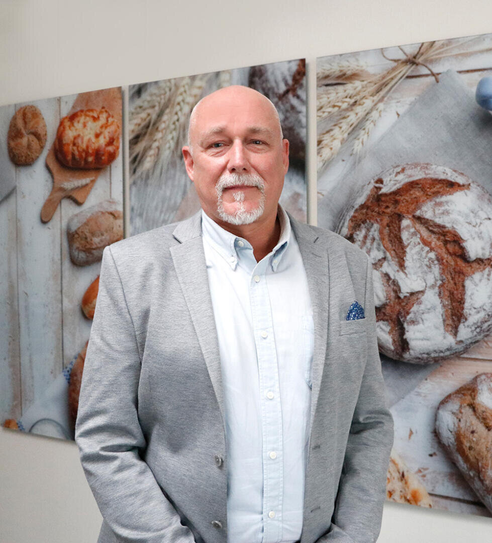 Ein Mann mit Glatze und weißem Spitzbart, der einen hellgrauen Blazer und ein weißes Hemd trägt, steht vor einem Wandbild mit Nahaufnahmen von Brot und Weizen.