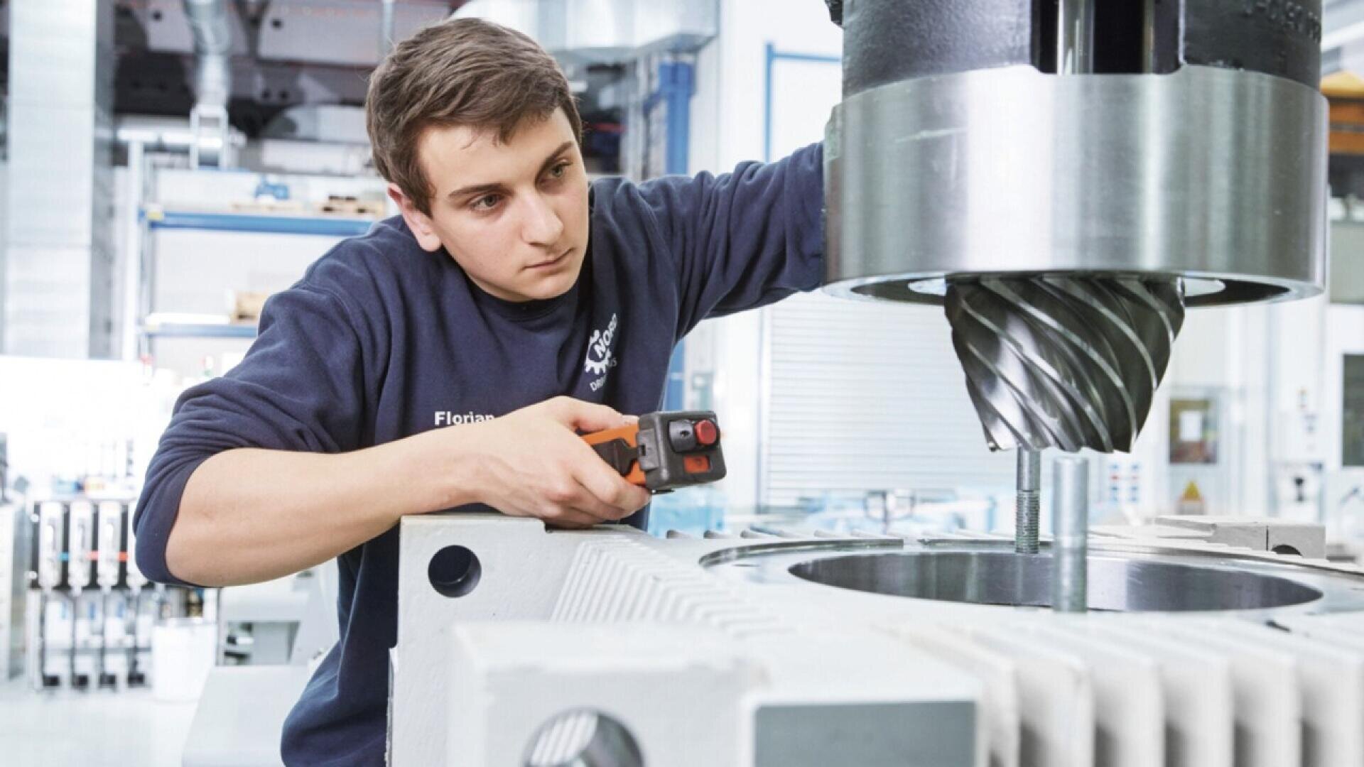 A young man in a workshop operates a handheld power tool, working on a large industrial machine with a metal drill or milling head. The environment appears modern and well-lit.