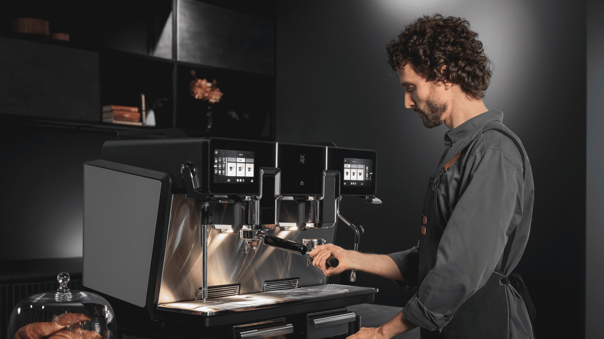 Ein Barista in dunklem Hemd und Schürze bedient eine moderne Espressomaschine mit zwei Köpfen in einem eleganten, schwach beleuchteten Café-Ambiente.