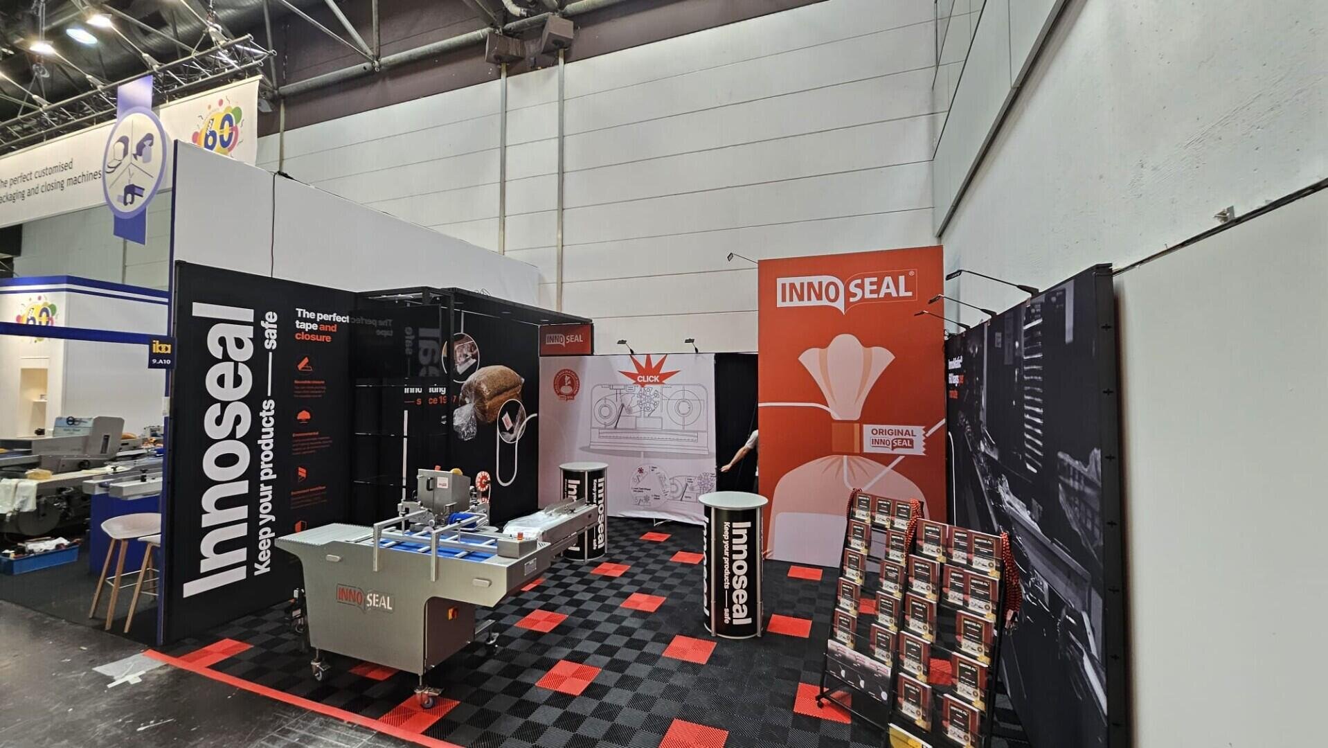 Ein Messestand für Innoseal mit Versiegelungsmaschinen, Produktdisplays, Informationsbannern und Broschüren auf einem schwarz-rot karierten Teppich. Der Stand ist mit Markenschildern und übersichtlich angeordneten Werbematerialien ausgestattet.