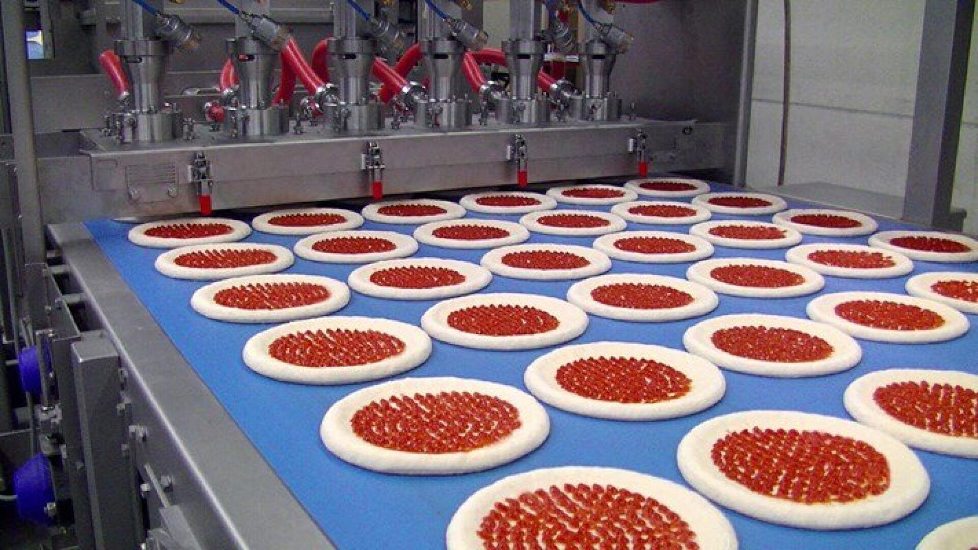 Reihen von rohen Pizzen mit Soße sind auf einem blauen Förderband angeordnet und laufen durch eine automatische Maschine, die die Soße in präzisen Kreisen auf jeden Teigboden in einer Lebensmittelproduktionsanlage verteilt.