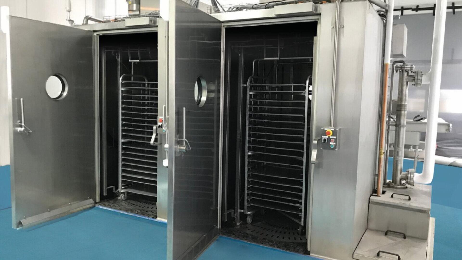 Two large stainless steel industrial ovens with open doors, revealing empty racks inside, are set in a clean facility with blue flooring and visible control panels on the right side.
