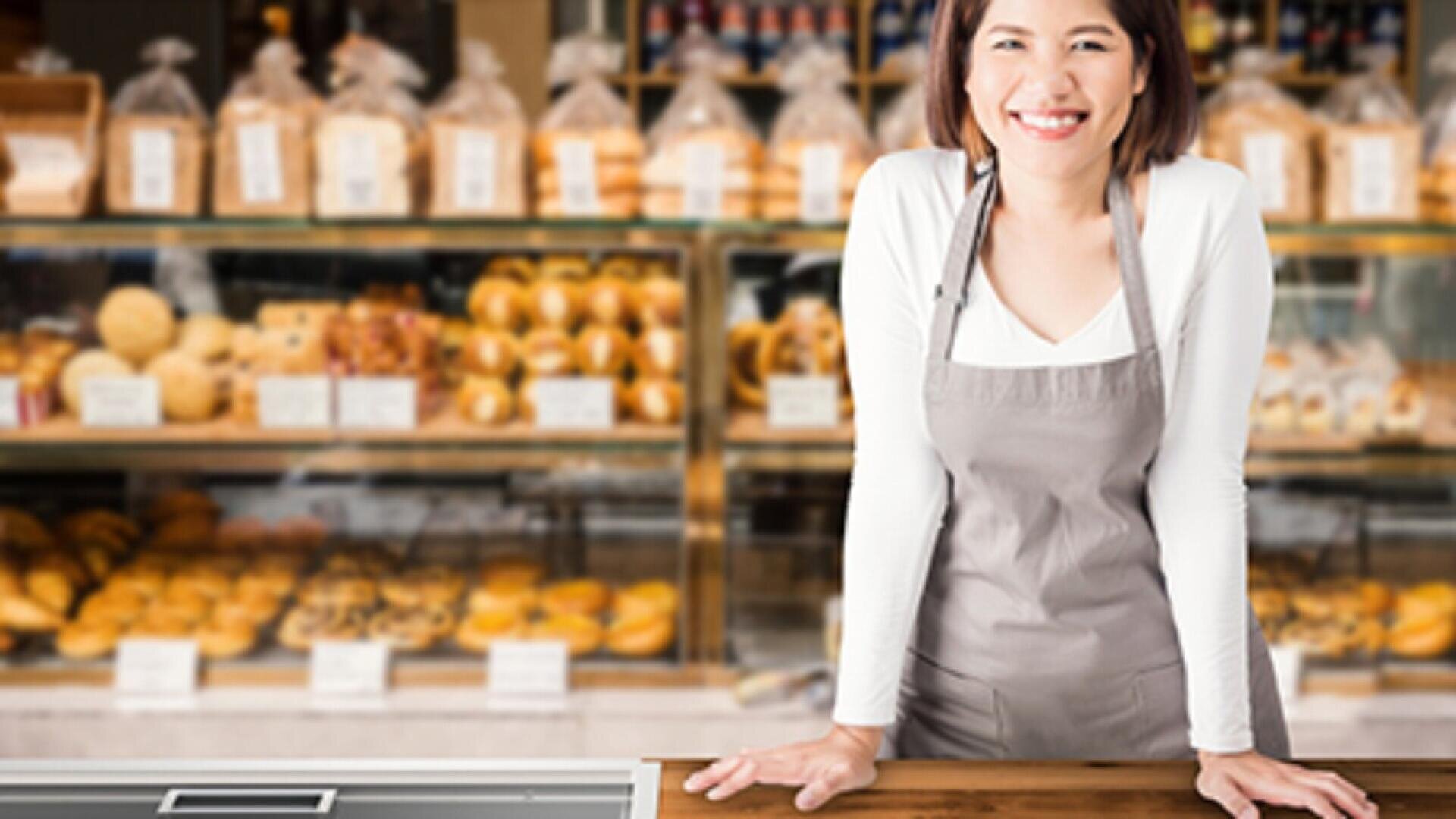 A smiling woman in an apron stands behind a wooden counter in a bakery, with shelves of bread and pastries displayed in the background.