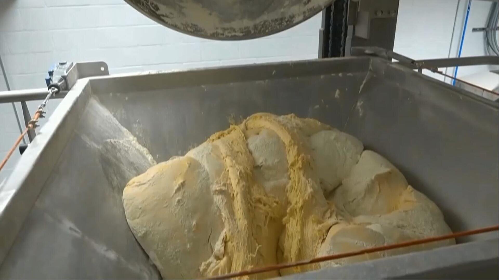 A large industrial mixer holds a batch of bread dough, partially mixed, inside a metal container in a commercial bakery setting.