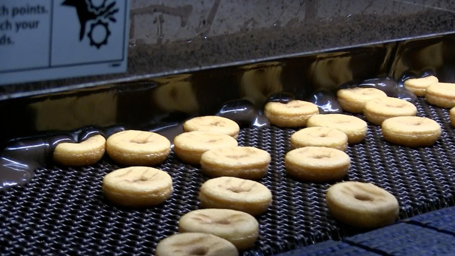Rows of donuts are moving along a metal conveyor belt inside a factory, likely during the frying process. A warning sign with an icon is visible in the upper left corner.