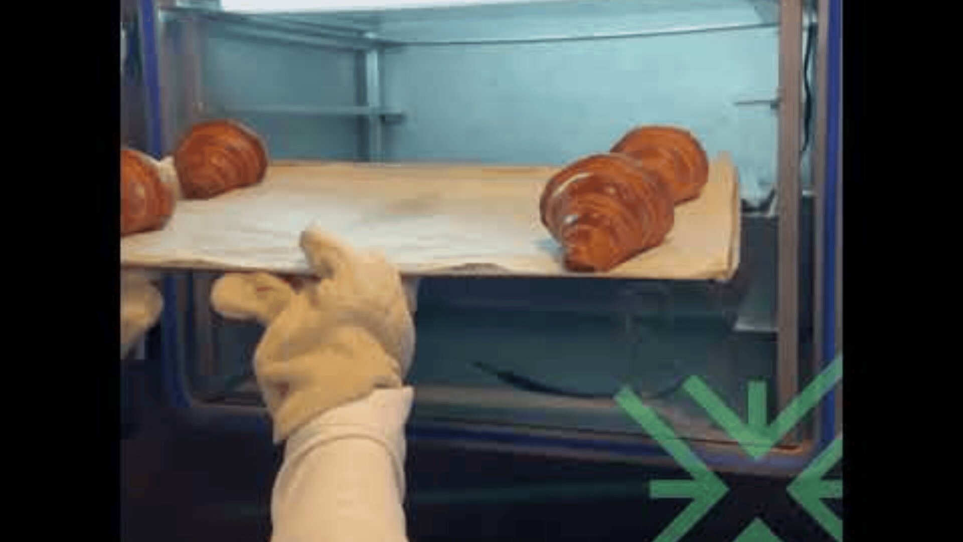 A person wearing white oven mitts is placing a baking tray with croissants into an oven. The tray has parchment paper, and three croissants are visible. A green geometric logo is in the bottom right corner.