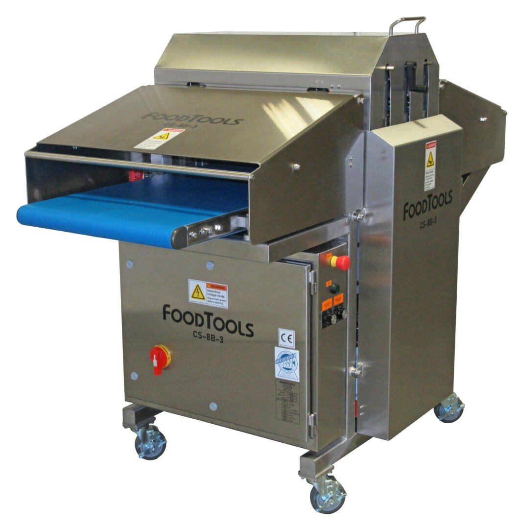 A stainless steel industrial food slicing machine labeled FOODTOOLS, with a blue conveyor belt, control buttons, safety warning labels, and wheels for mobility.