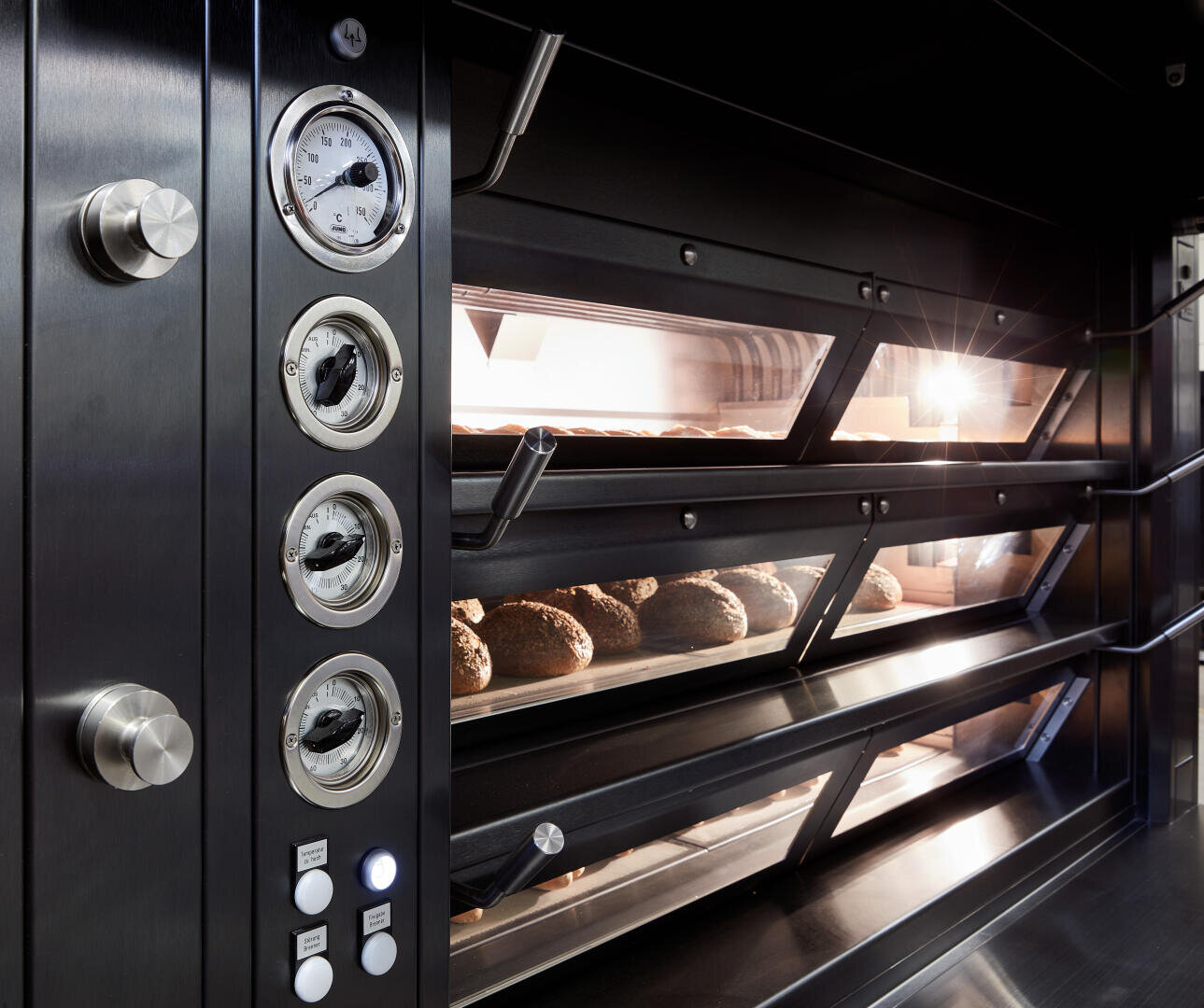 Nahaufnahme eines kommerziellen Brotofens mit Drehreglern und Messinstrumenten auf der linken Seite und mehreren Brotlaiben, die durch die Glasfenster auf der rechten Seite gebacken werden.
