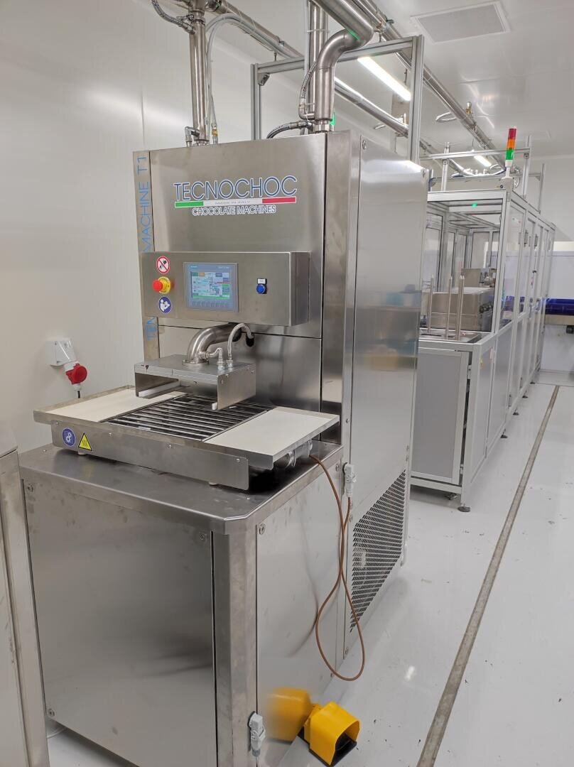 A stainless steel industrial machine labeled TECNOCHOC with a touchscreen control panel stands in a clean, well-lit facility. The machine has trays and pipes connected, and yellow foot pedals are on the floor in front.