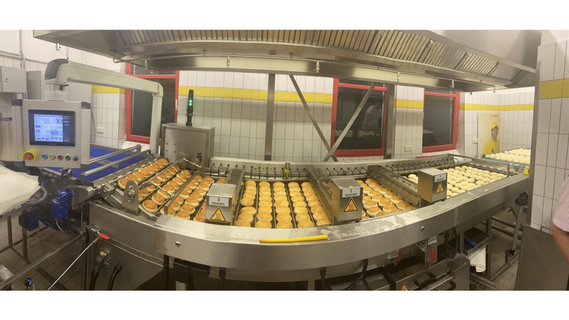 A production line in a bakery factory with rows of round pastries or buns moving along conveyor belts, surrounded by stainless steel machinery and tiled walls. A control panel is visible on the left.