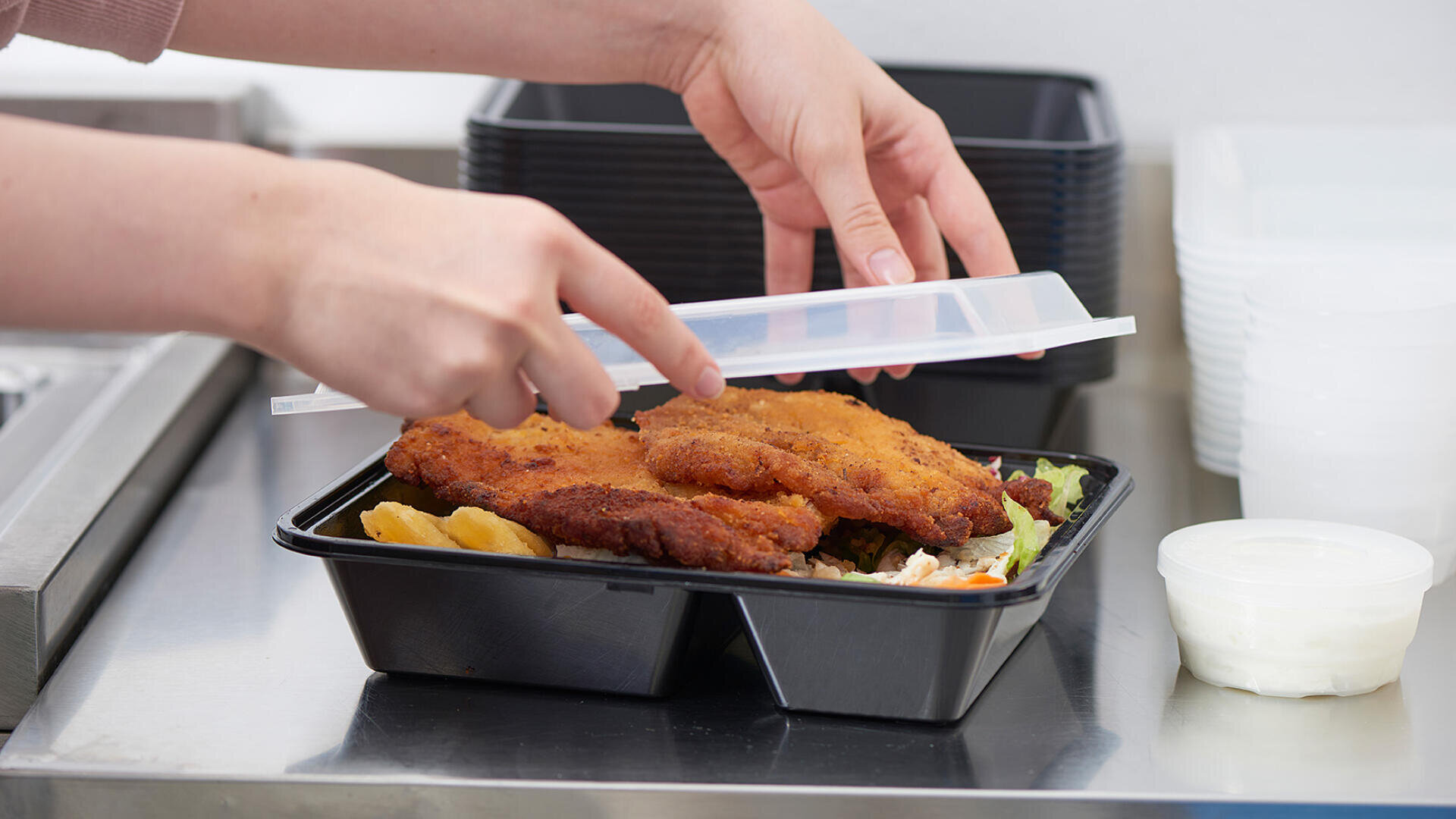 Eine Person verschließt einen Plastikbehälter mit panierten Schnitzeln, Kartoffelecken und Gemüse. Der Behälter steht auf einer Metallfläche. Daneben stehen leere schwarze Behälter und ein kleiner Becher mit Soße.