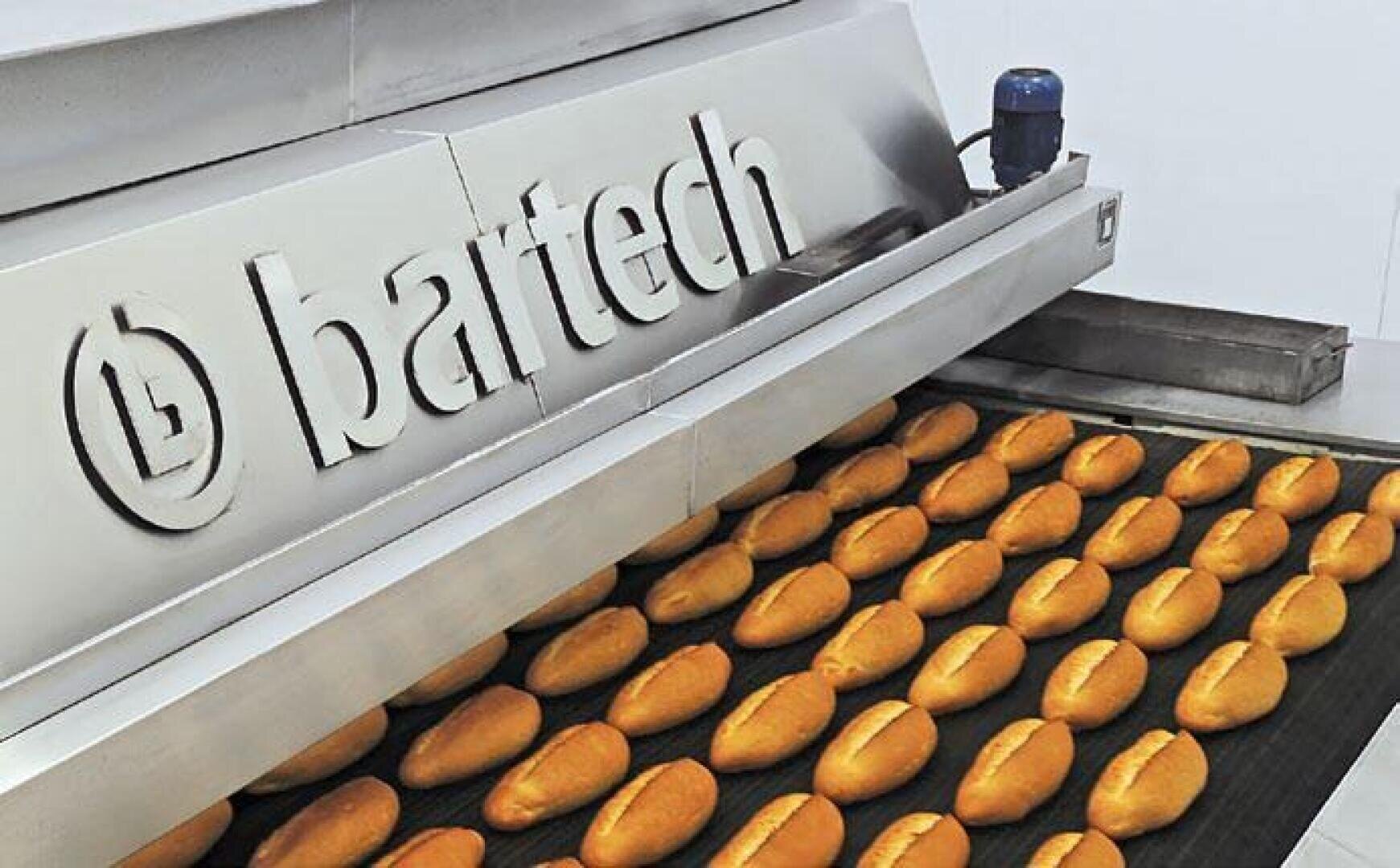 In a modern bakery, golden-brown bread rolls run along a conveyor belt under a stainless steel machine labeled "bartech".