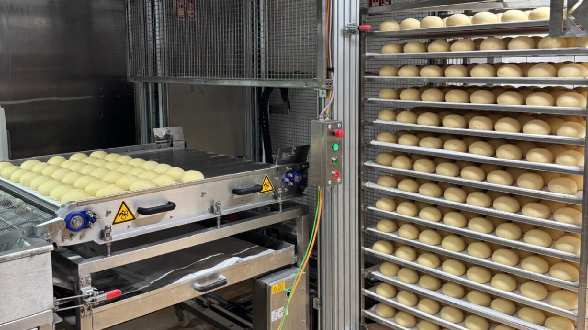 Rows of dough balls are lined up on metal trays and racks in an industrial bakery, with machinery and conveyor belts visible, indicating an automated bread or pastry production process.