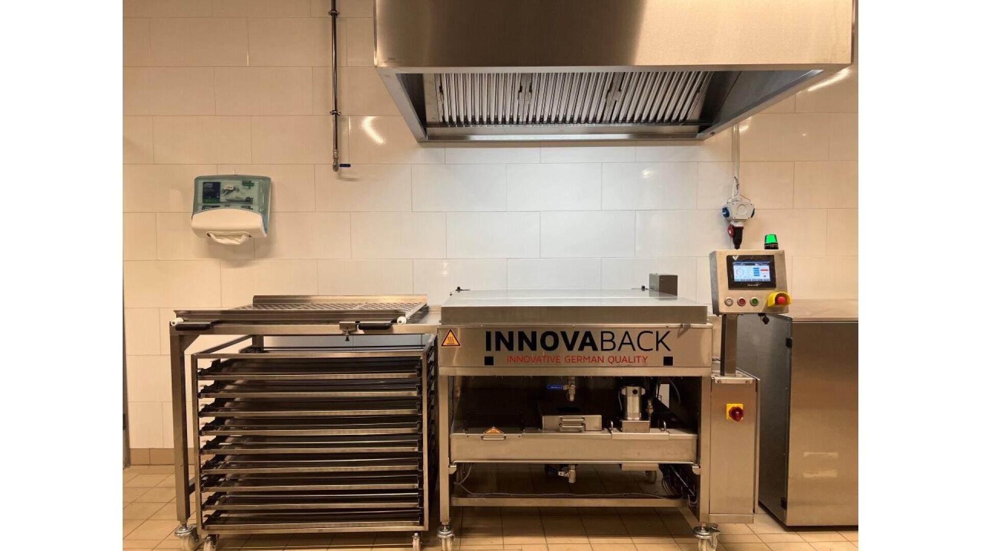 A commercial kitchen with a large stainless steel oven labeled INNOVABACK, a metal rack with trays, a wall-mounted dispenser, and control panel, all set against white tiled walls and a vent hood above.