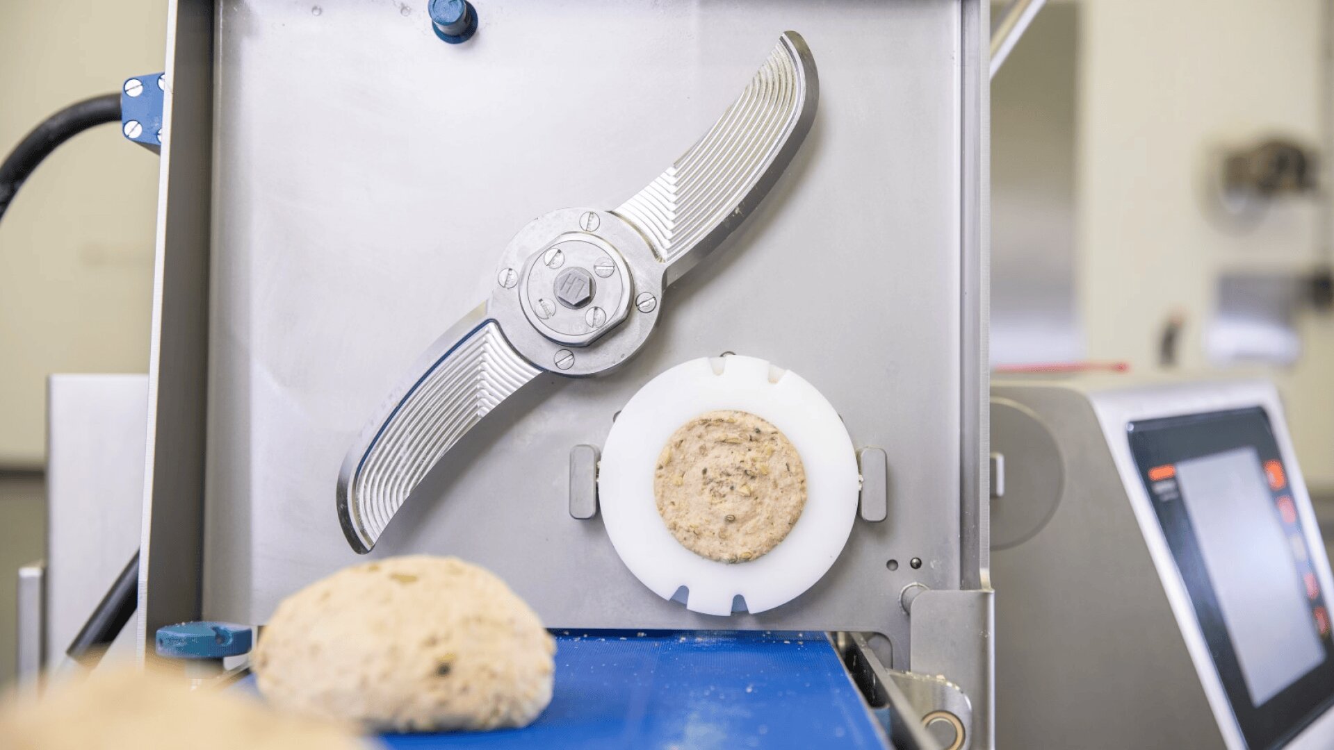 Close-up of an industrial food processing machine with a large metal blade and round mold shaping dough on a conveyor belt. A screen and another part of the machine are visible to the right.