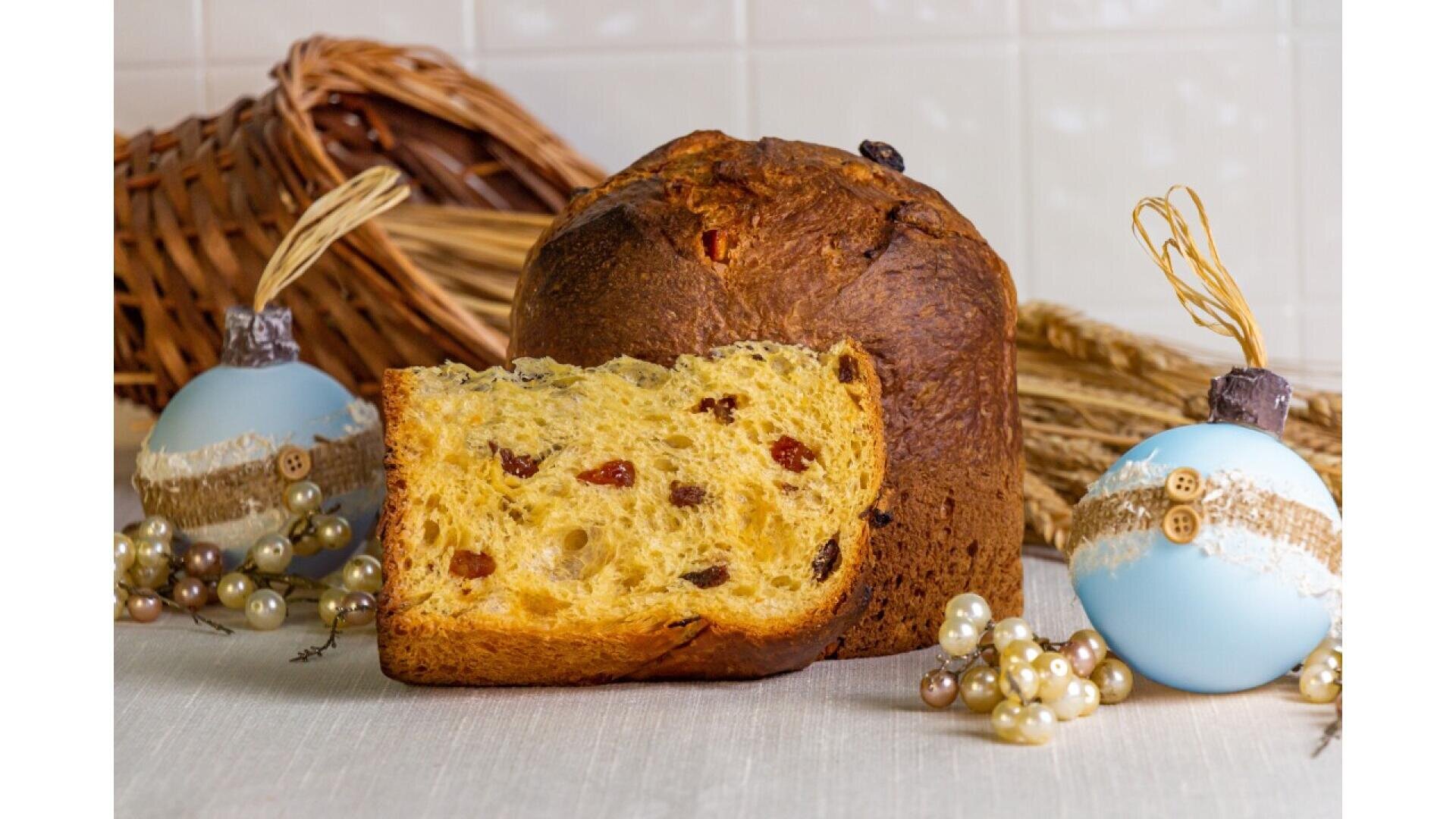 Ein aufgeschnittener Panettone mit sichtbaren Trockenfrüchten liegt auf einem Tisch, umgeben von dekorativen blauen Ornamenten, Perlengirlanden und rustikalem Weidengeflecht im Hintergrund.