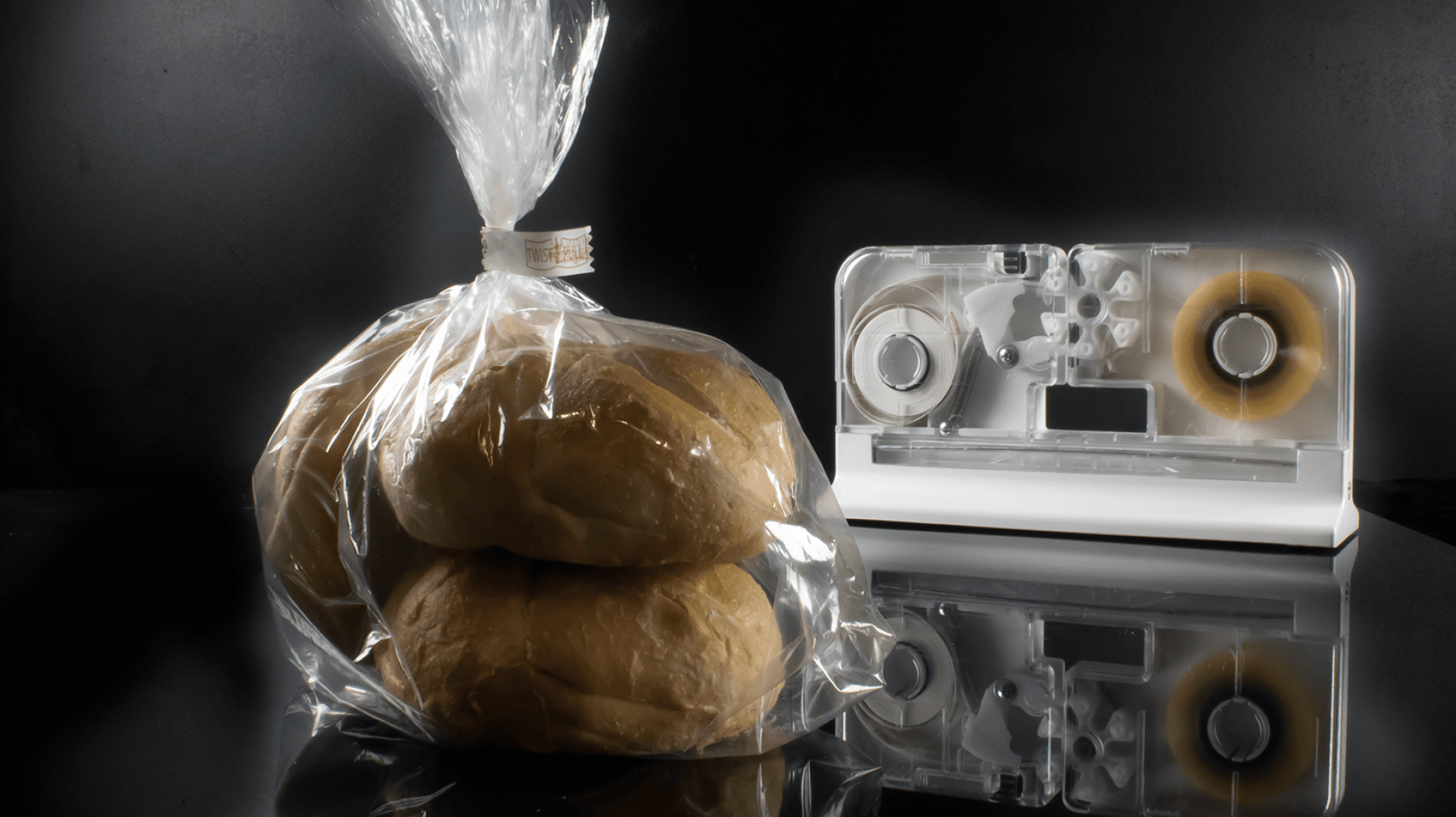 A bag of bread rolls sealed with a plastic twist tie sits on a reflective surface next to a bag sealing tape dispenser against a dark background.