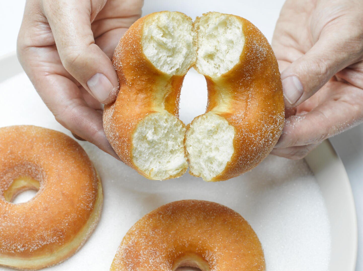 Close-up of hands breaking a sugar-coated donut in half over a plate with two whole donuts. The fluffy, soft texture of the donut’s inside is visible.